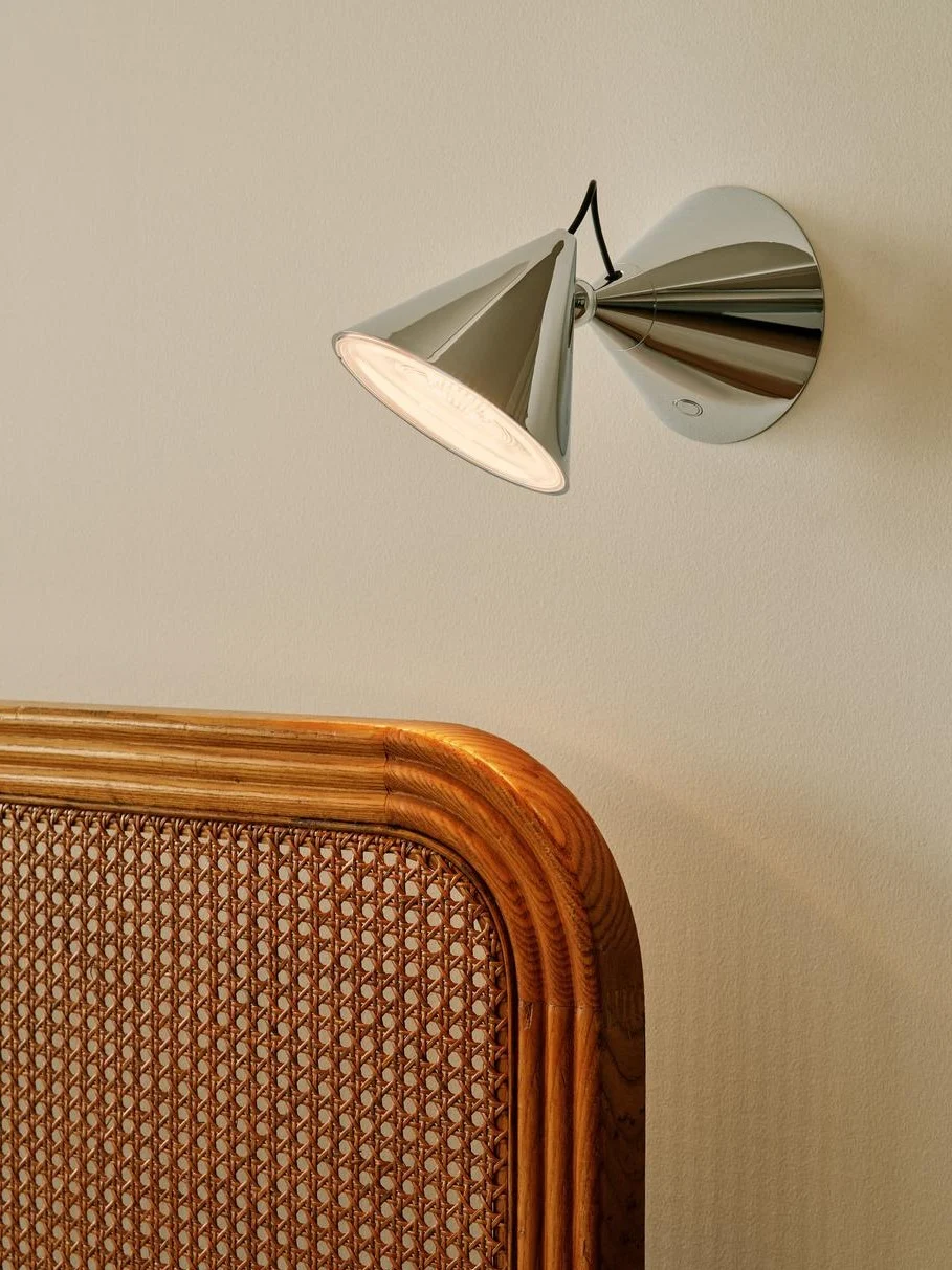 Moderne krom vegglampe med konisk skjerm som lyser opp en lys vegg over en flettet rotting- og sengegavl av tre.