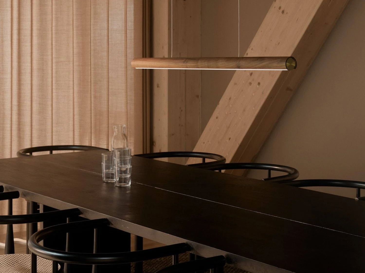 Et moderne spisestue med et langt mørkt trebord, svarte stoler og en elegant pendellampe i tre. En glasskaraffel og tre glass står på bordet.