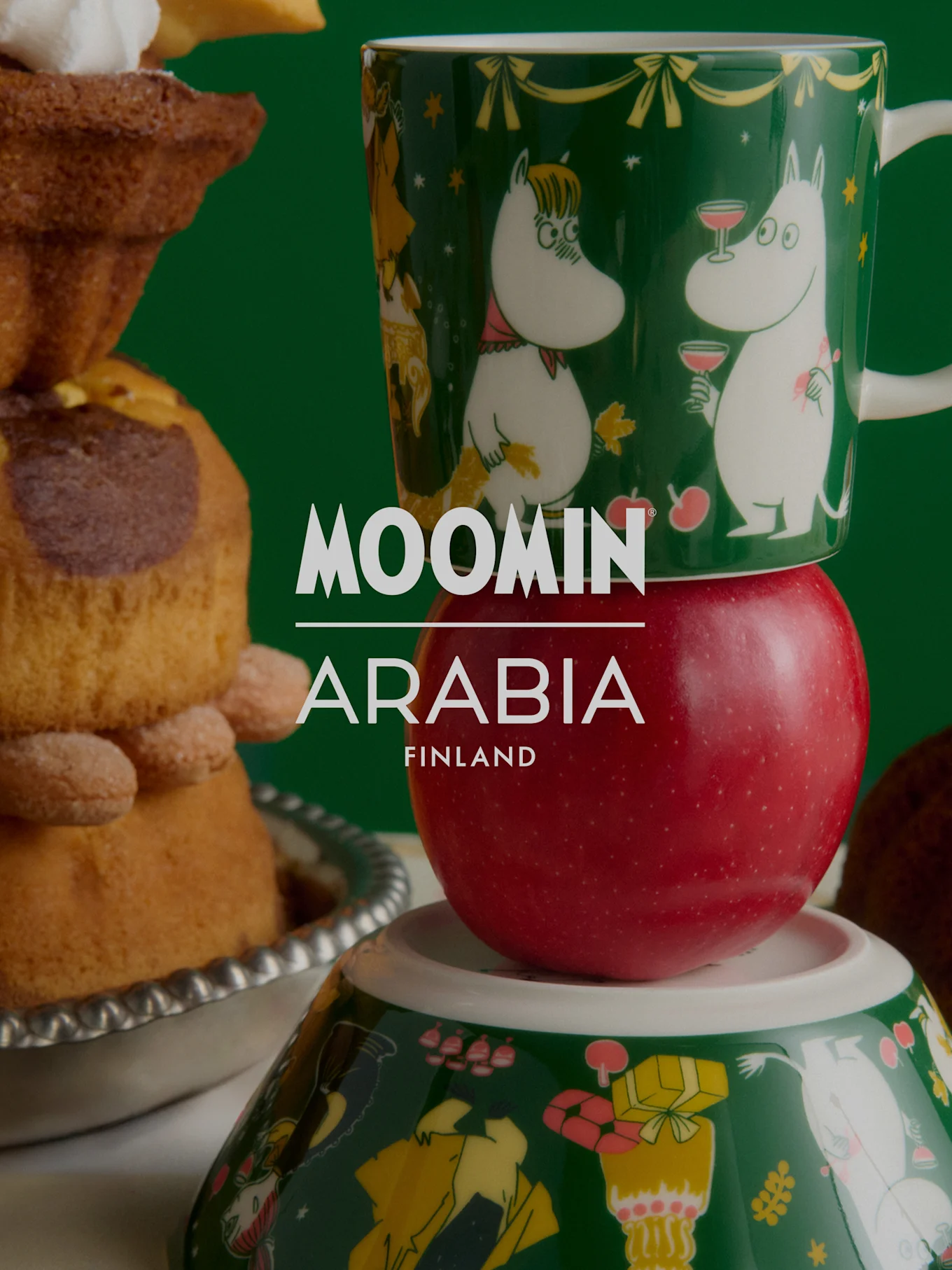 Moomin Arabia julekolleksjon: Et grønt Mummi-krus på et rødt eple, oppå en Mummi-skål, ved siden av festlige bakervarer.