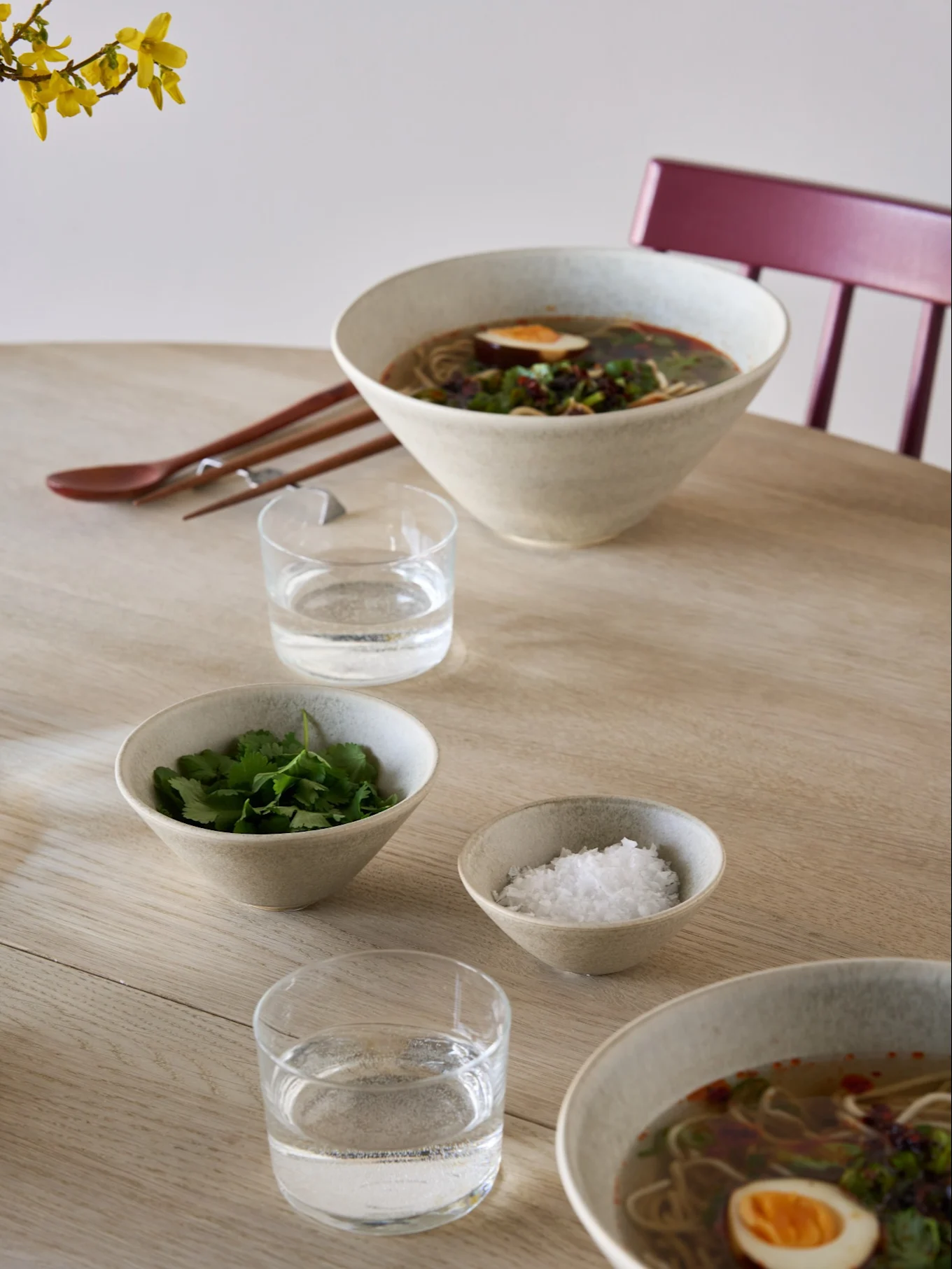 To boller ramen med egg og urter, sideboller med koriander og salt, vann, spisepinner og en skje på et lyst trebord.