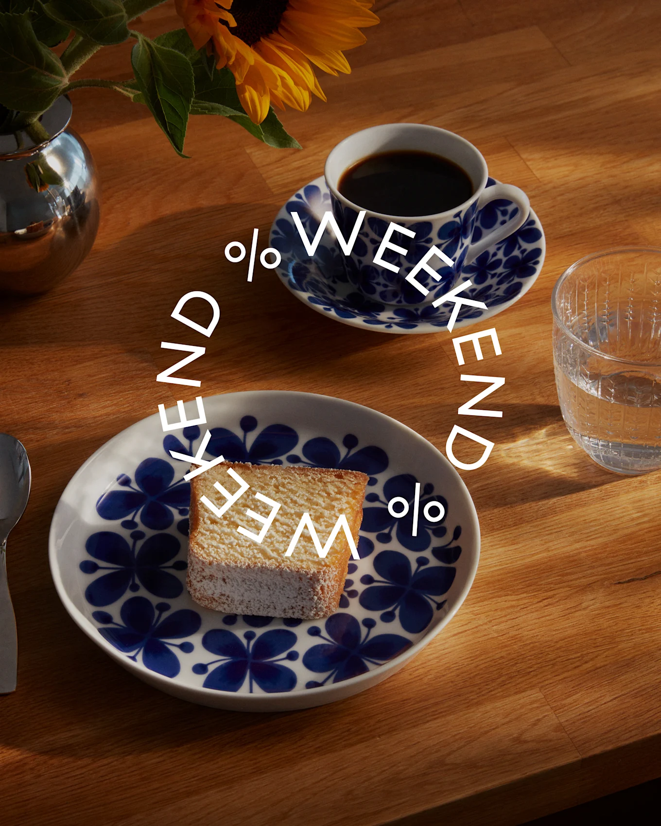 Et kakestykke med melis på et hvitt fat med blått blomstermønster, kaffe i kopp, vannglass og solsikke i vase på trebord. Tekst: Weekend %