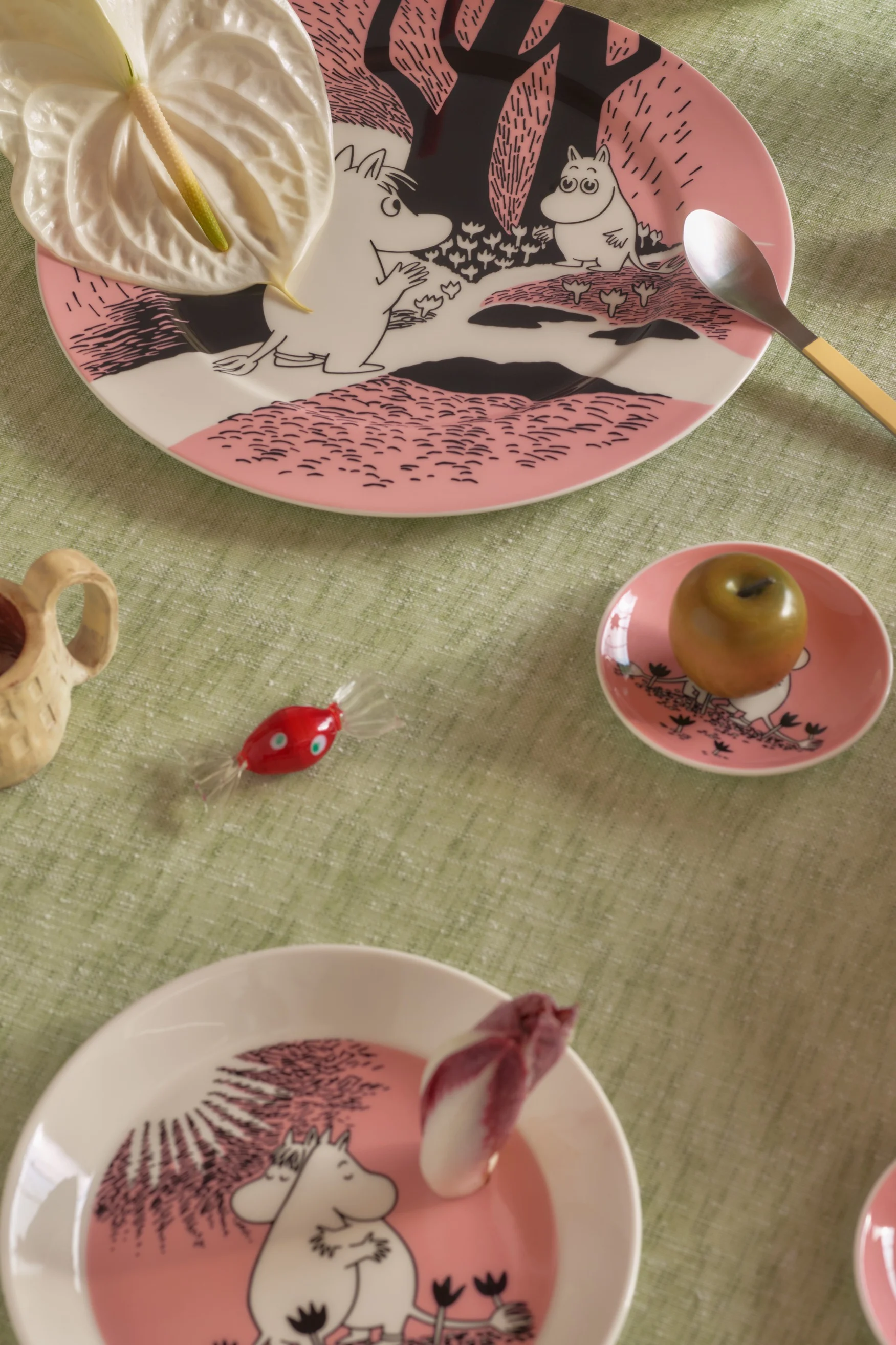 Bord dekket med rosa Mummi-tallerkener, anthuriumblomst, skje, grønt eple, rødt godteri og en trekopp på en grønn duk.