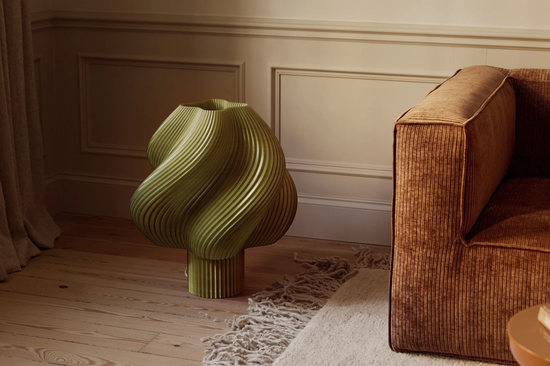 En olivengrønn, skulpturell, riflet gjenstand på et tregulv ved siden av en brun kordfløyelsofa og et teksturert teppe i et rom med profilerte vegger.