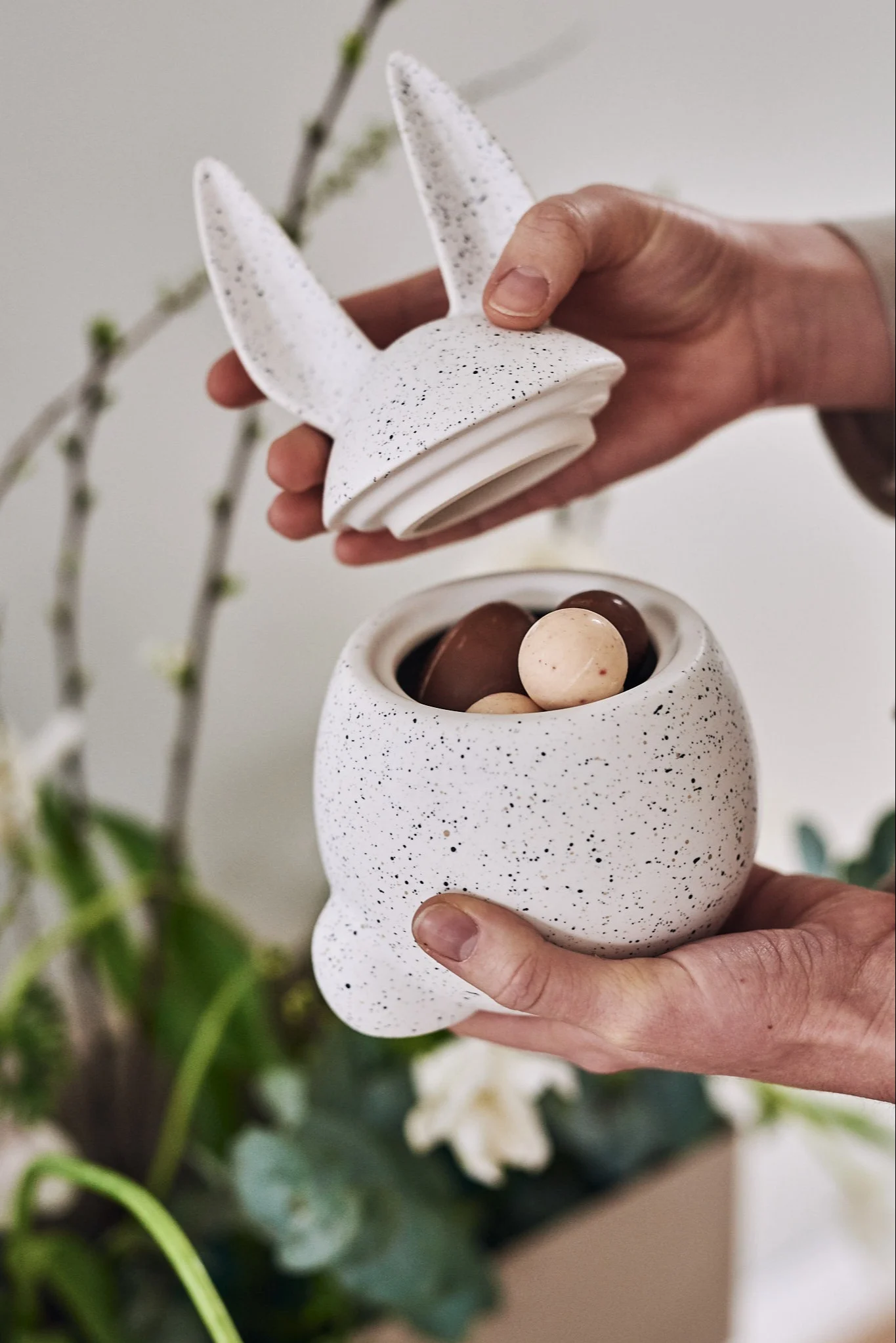 Hender åpner en hvit, flekkete kaninbeholder fylt med sjokoladepåskeegg.