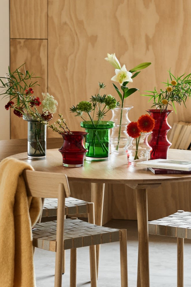 En samling Kosta Boda vaser i klart, rødt og grønt glass står på et spisebord med snittblomster.
