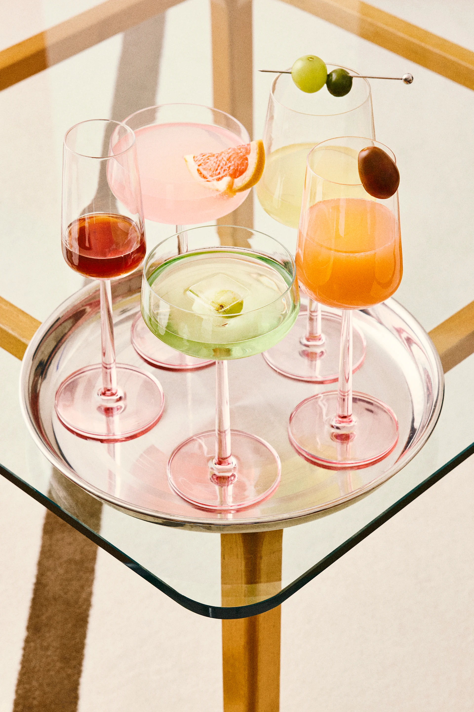 Fem fargerike cocktails på et sølvbrett, inkludert rosa, grønne, oransje og røde drinker med forskjellige garnityrer, på et glassbord.
