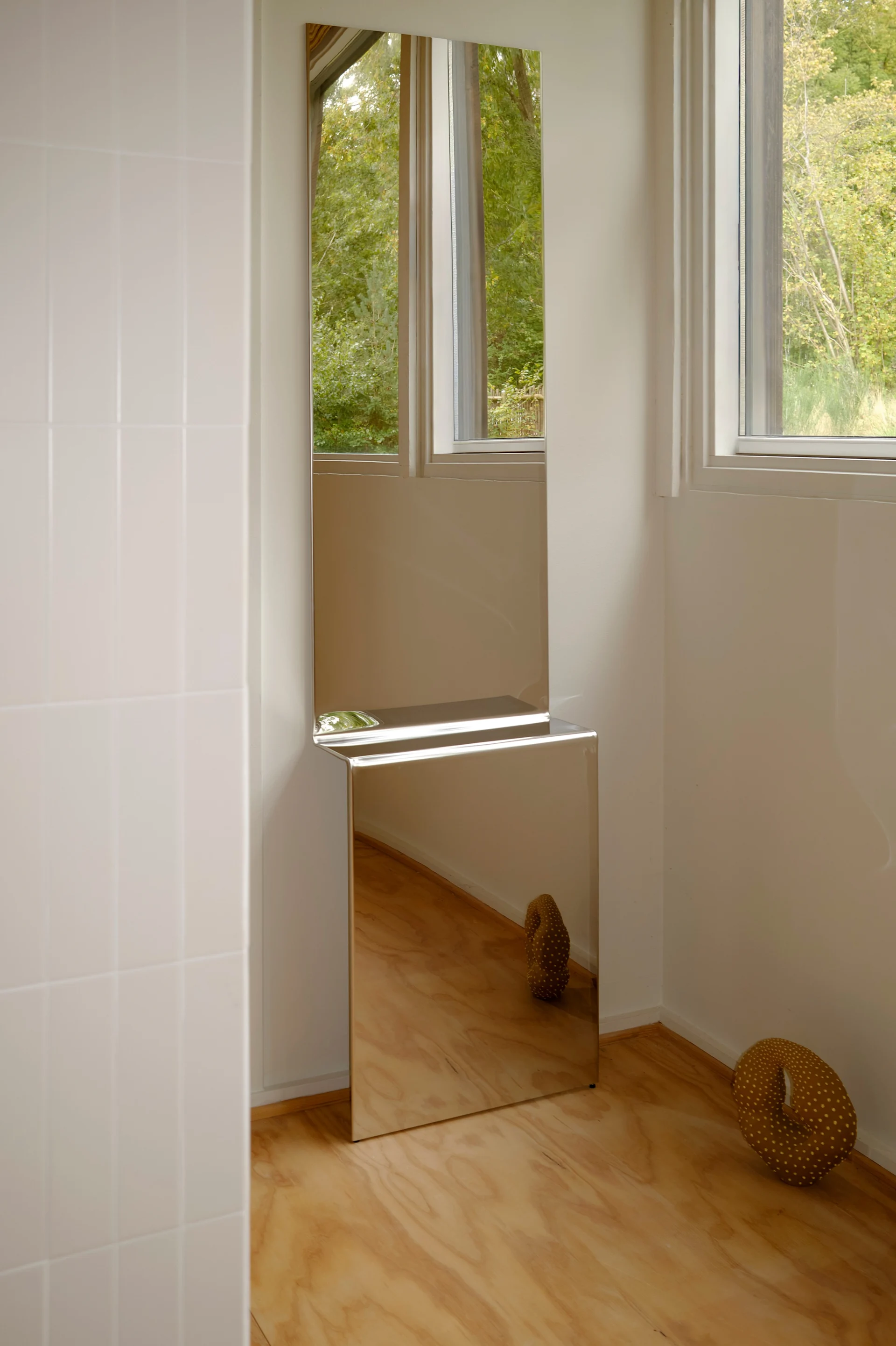 Høye, stablede speil i et moderne rom. Toppen reflekterer vindu og trær; bunnen reflekterer et tregulv og en brun prikkete skulptur.