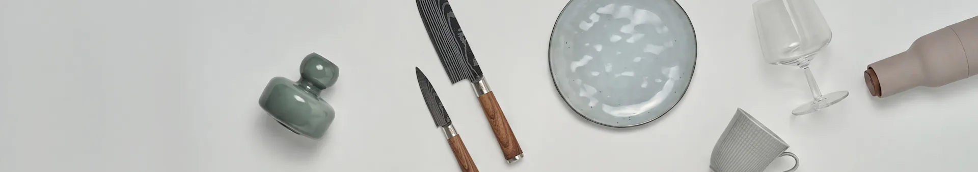 Ulike kjøkken- og spisevarer, inkludert en grønn vase, to kniver, en flekket tallerken, et vinglass, en beige flaske og et mønstret krus.