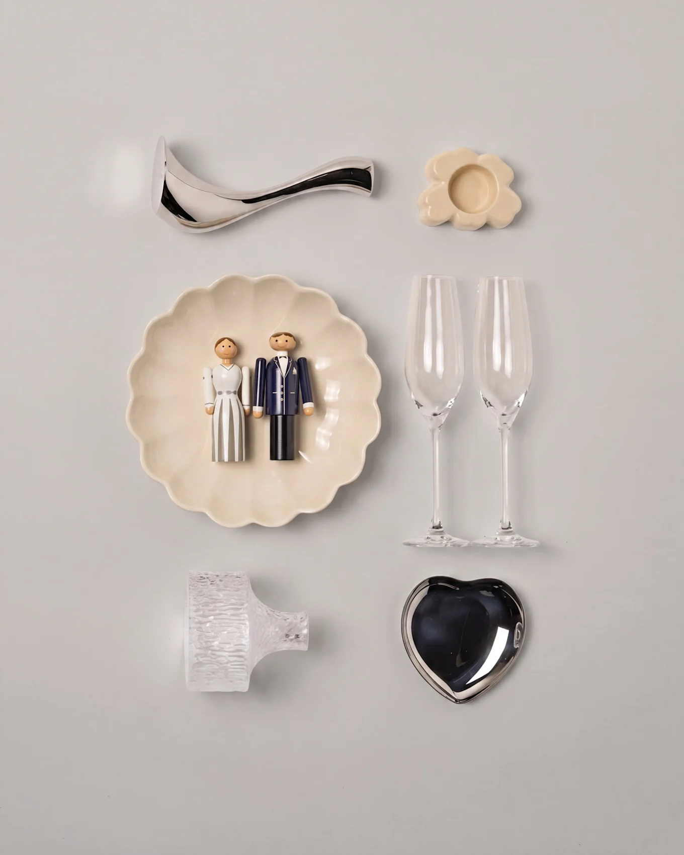 Bryllups-flat lay med trefigurer av brudepar på en muslingskål, to champagneglass, et sølvfarget hjertefat og diverse dekorasjoner.
