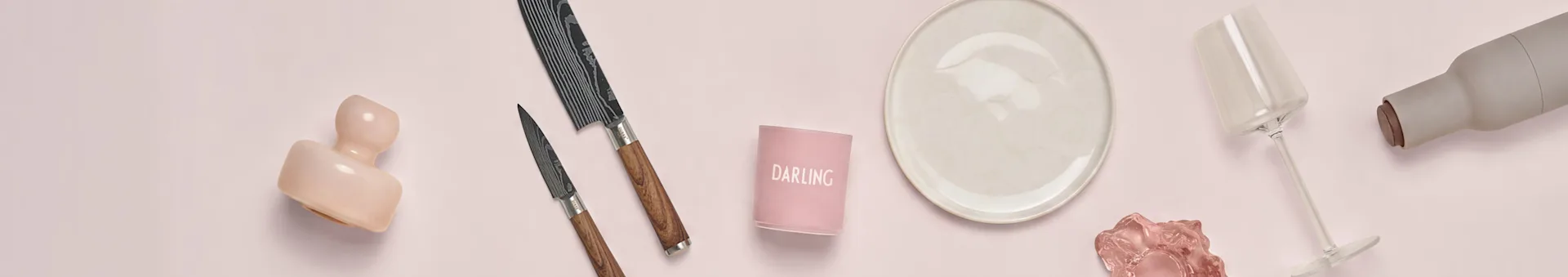 Utvalg av kjøkken- og spiseartikler på rosa bakgrunn: en rosa skulptur, to mønstrede kniver, en "DARLING" lysestake, en kremfarget tallerken, et klart vinglass og en grå flaske.