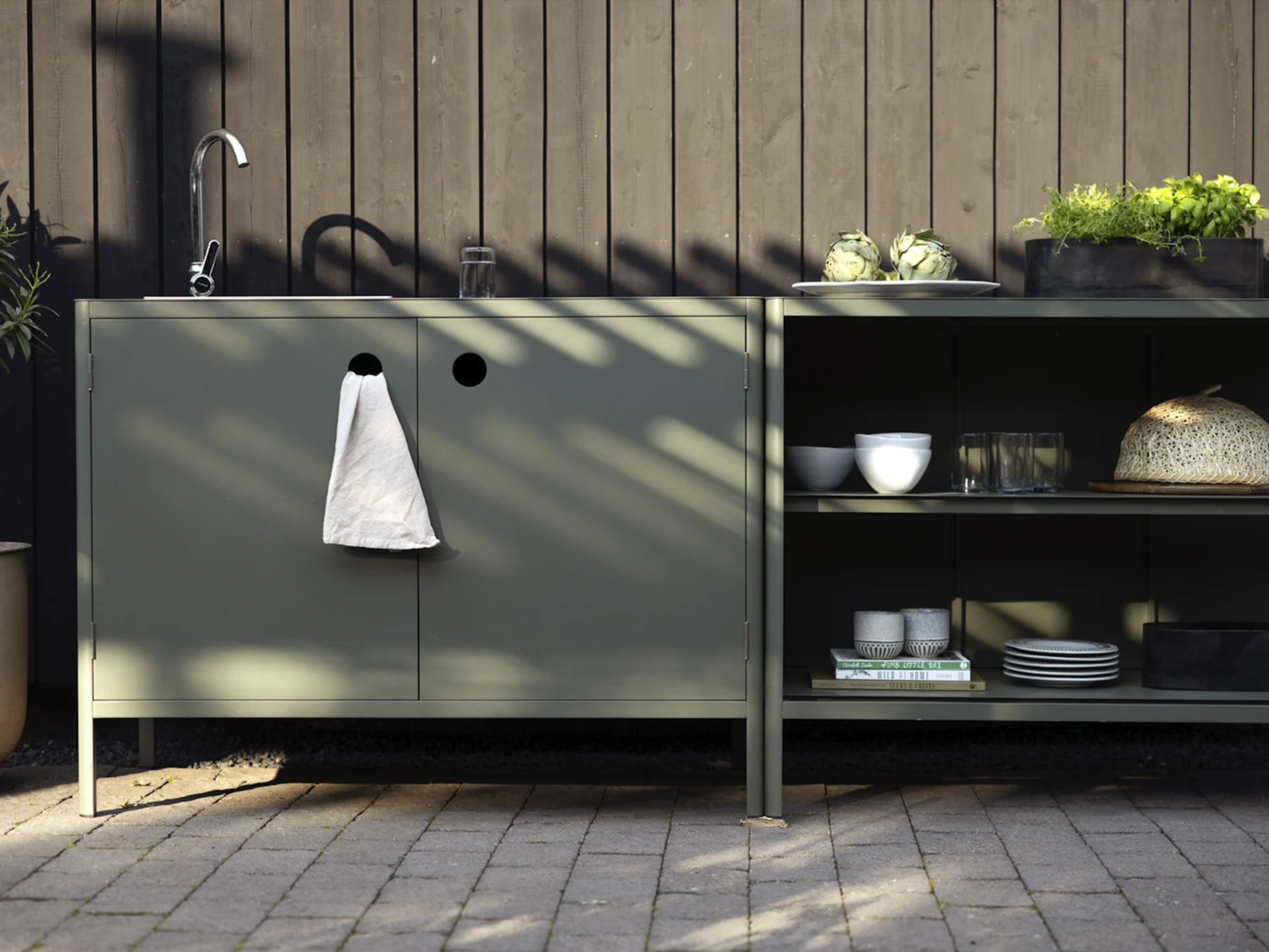 Utekjøkken med mørkegrønne skap, vask, kran og hyller med artisjokker, urter og servise.