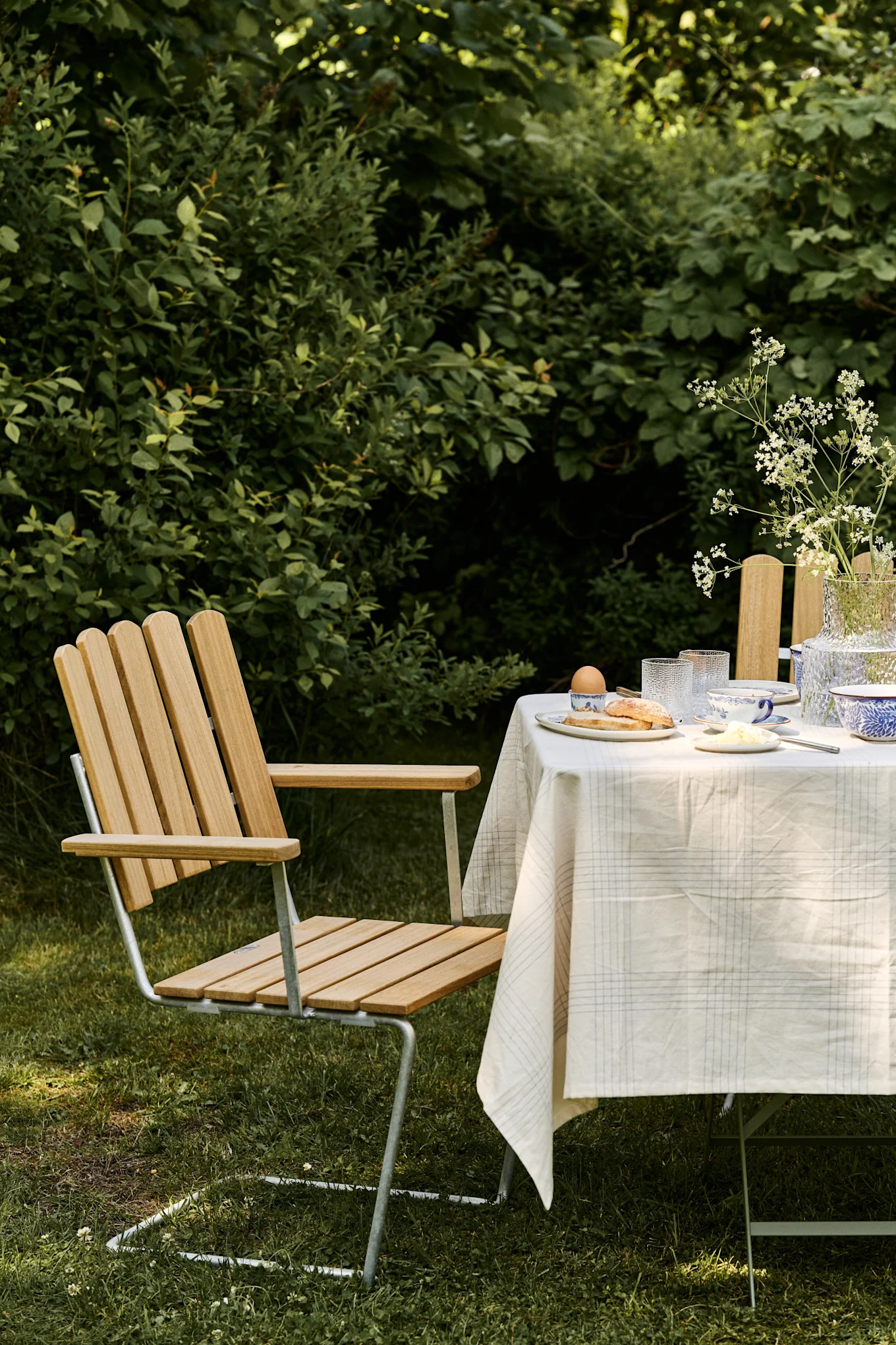 En frokostscene utendørs med en stol av lyst tre og metall ved et bord dekket med rutete duk, servise og en vase med hvite blomster.
