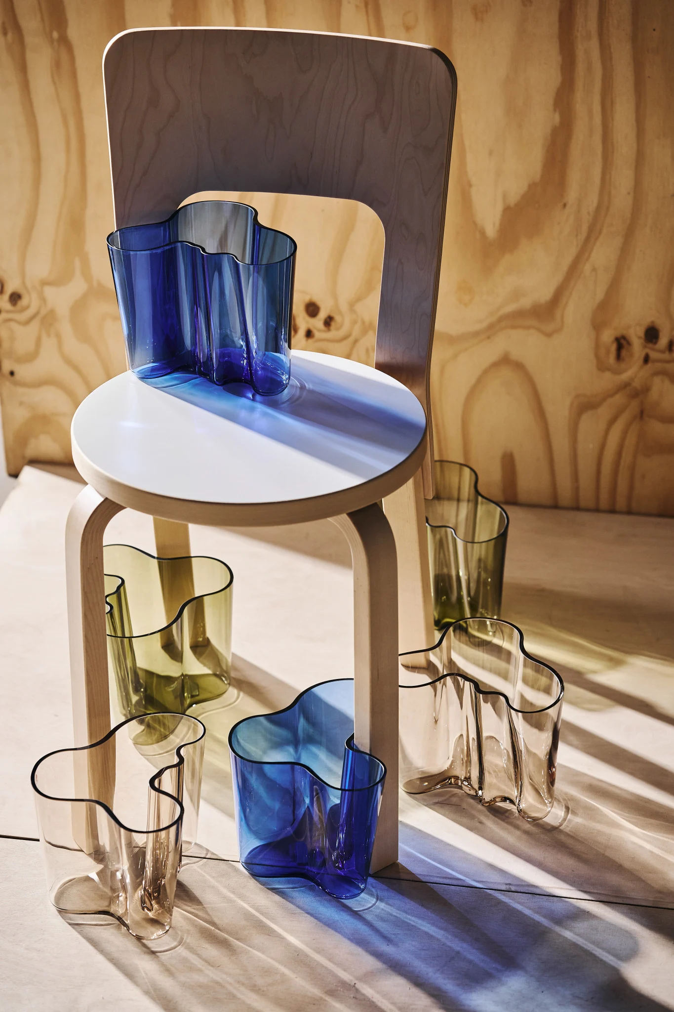 Flere bølgete glassvaser i blått, olivengrønt og klart, arrangert på og rundt en lys trepall med en trestol i bakgrunnen. Sollys kaster fargede skygger.