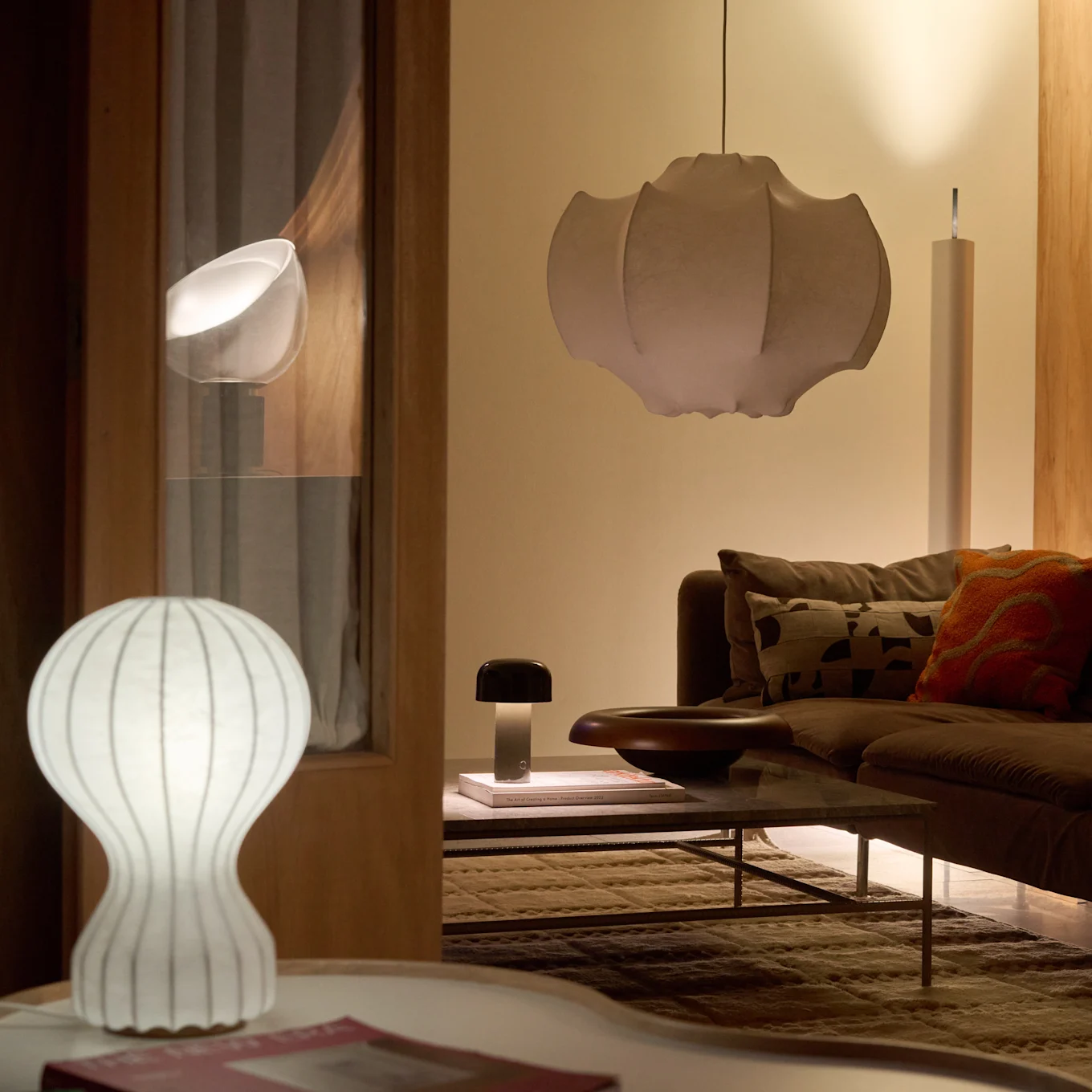 En stue med en brun sofa og salongbord, belyst av en hvit riflet bordlampe, en mørk sopplampe og en stor plissert pendellampe, som skaper en varm glød.