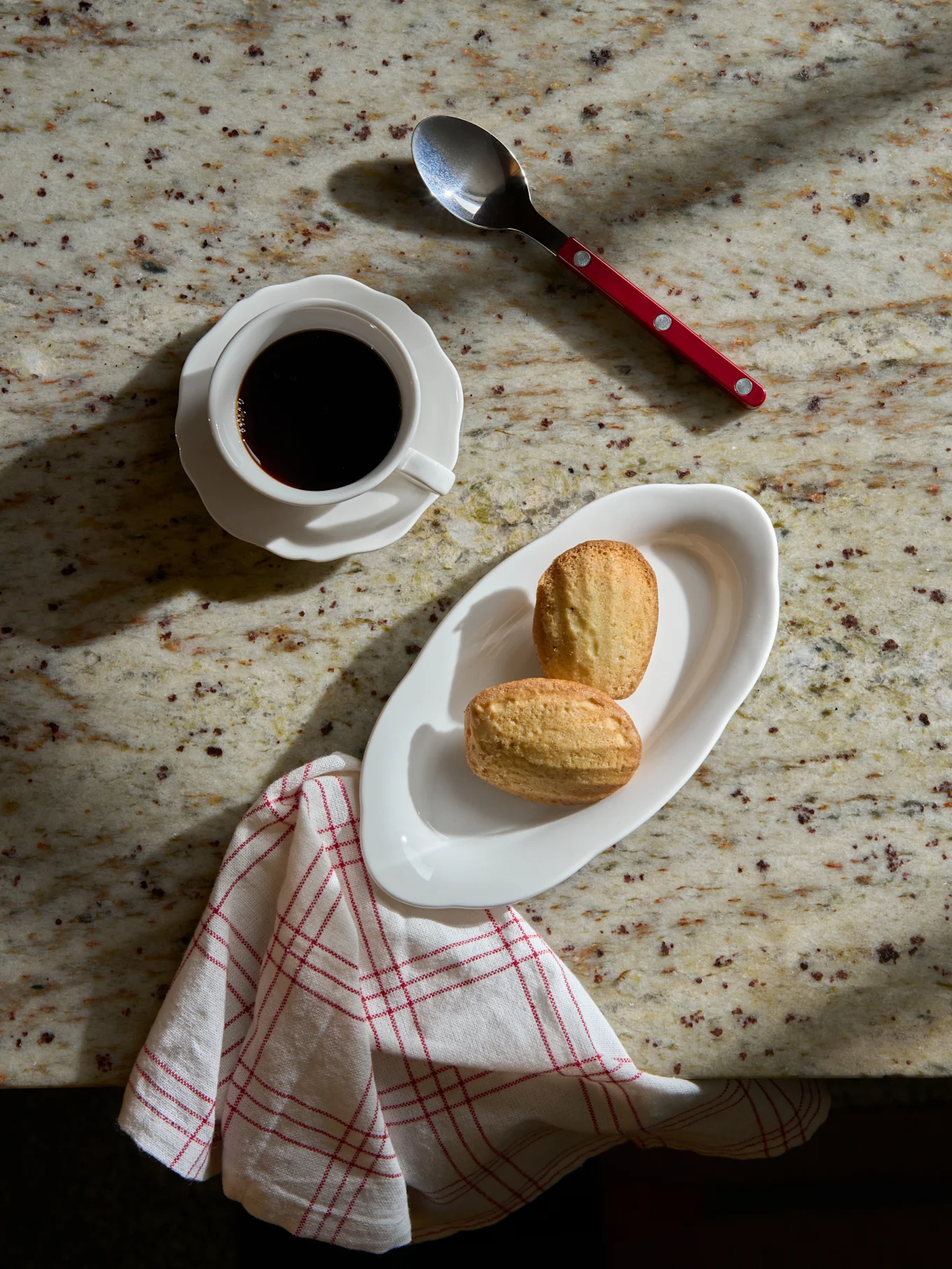 Ovenfra: Svart kaffe i hvit kopp, to madeleinekaker på hvit tallerken, skje og stripet serviett på marmorbenk.