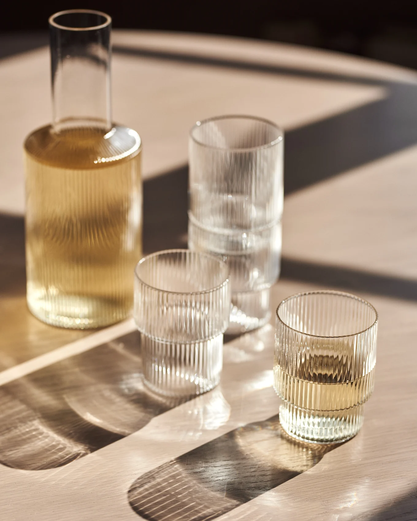 Glasskaraffel og riflede vannglass på et trebord med sollys.