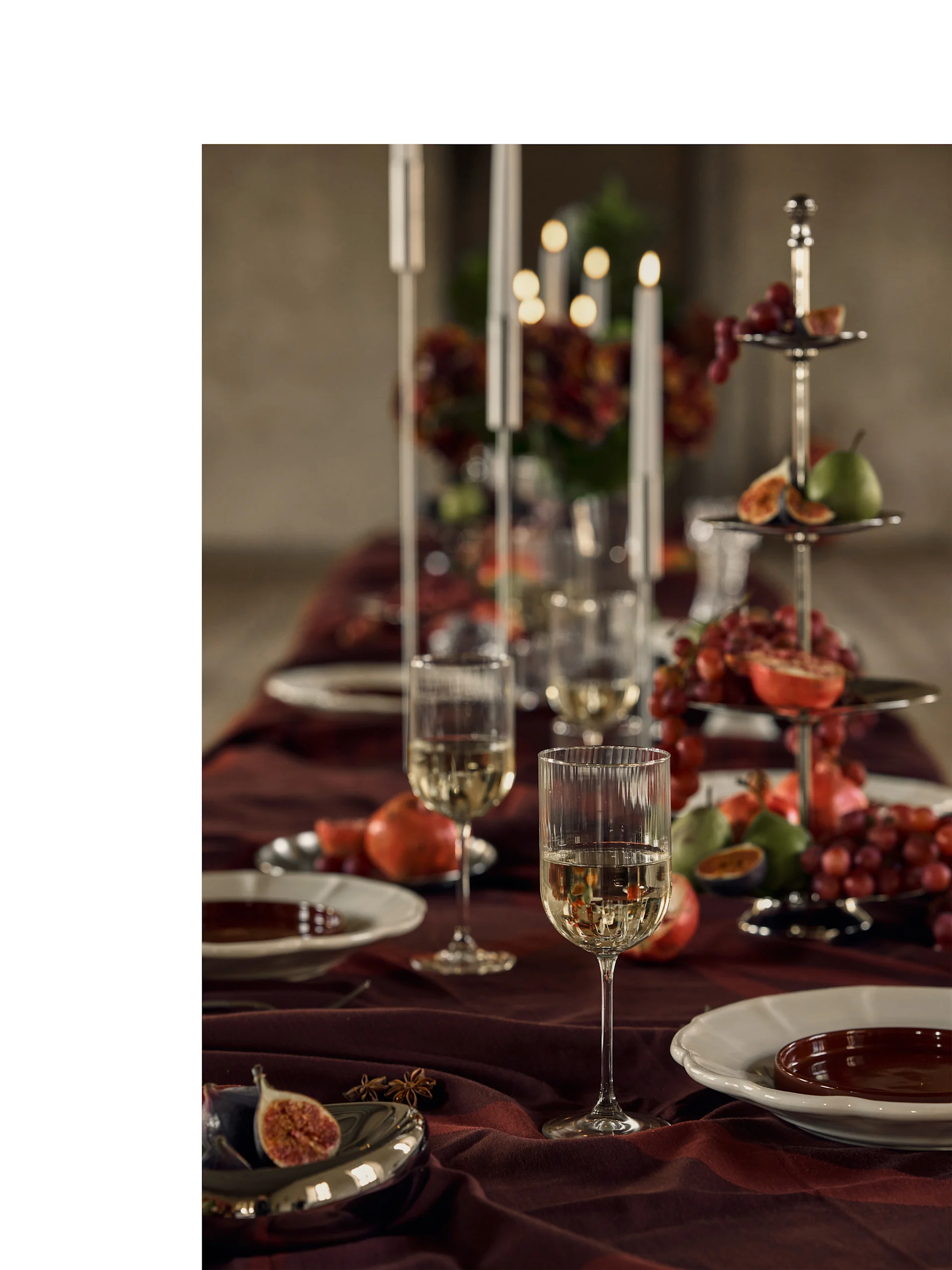 Festlig bord dekket med hvitvinsglass, tallerkener og etasjefat fylt med fargerike frukter og levende lys.