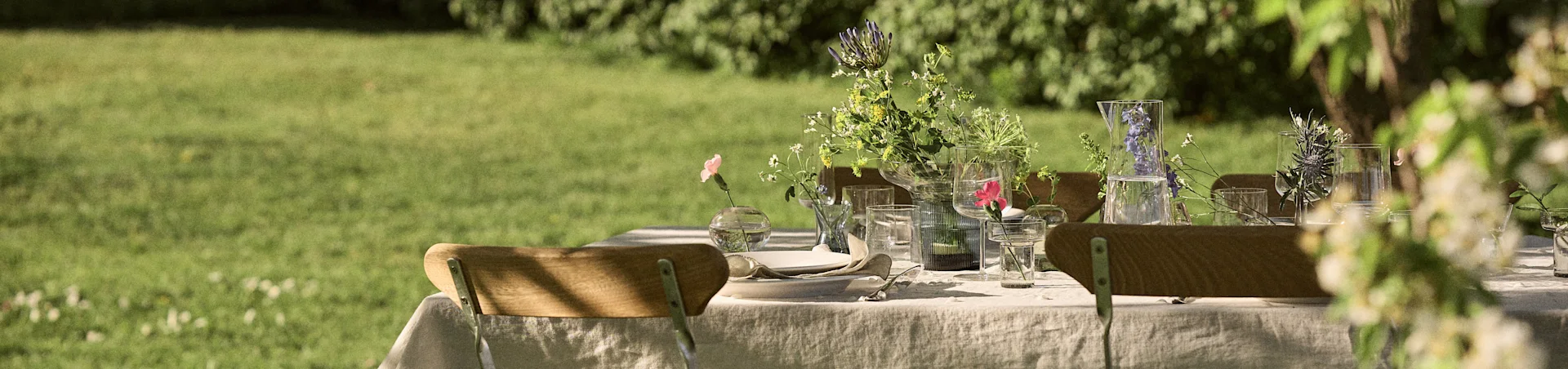 Et utendørsbord dekket med en hvit duk, tallerkener, bestikk og glassvaser med blomster, satt i en grønn hage.