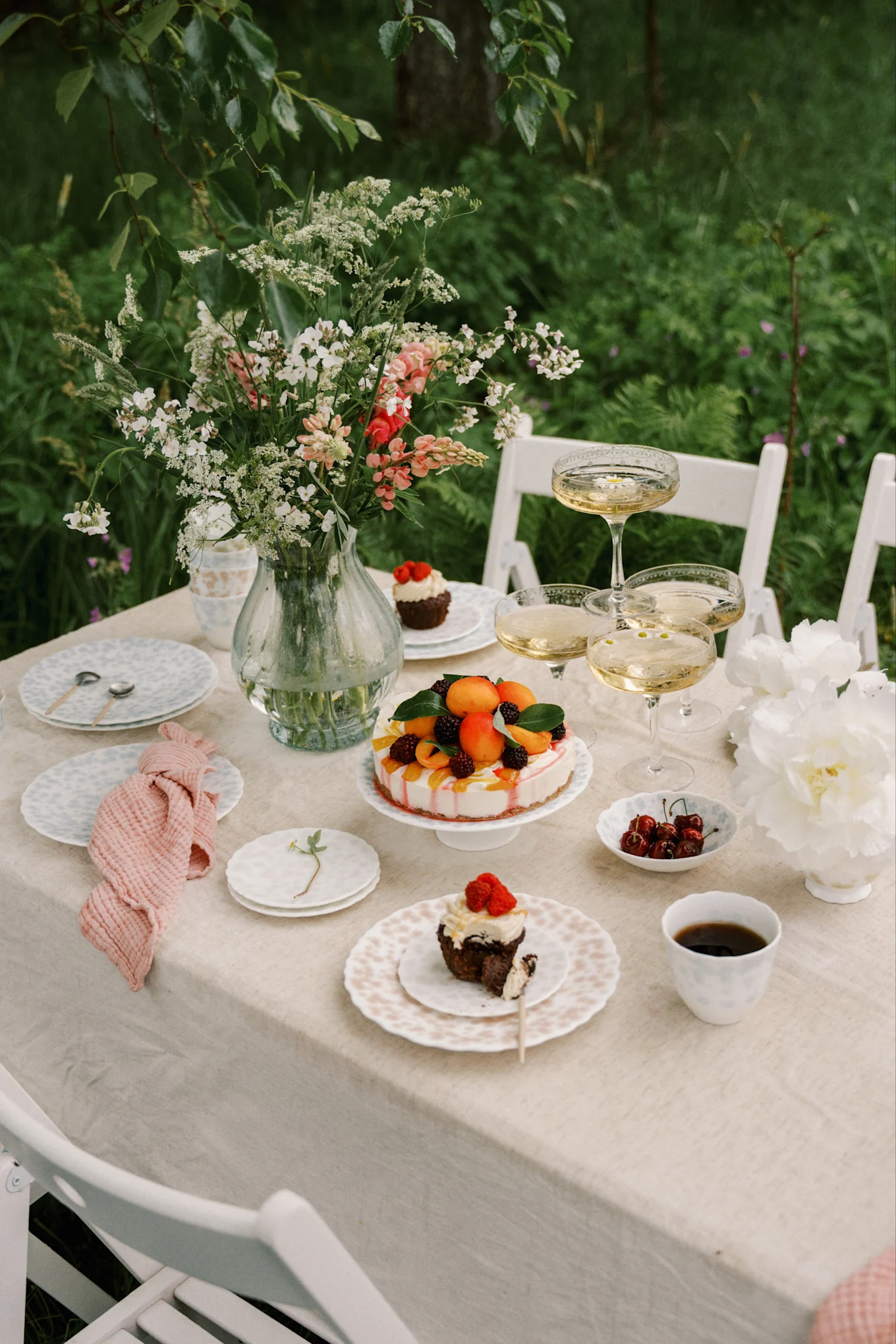 Utendørs bord dekket med blomstervase, champagne, fruktkake og cupcakes med bær.