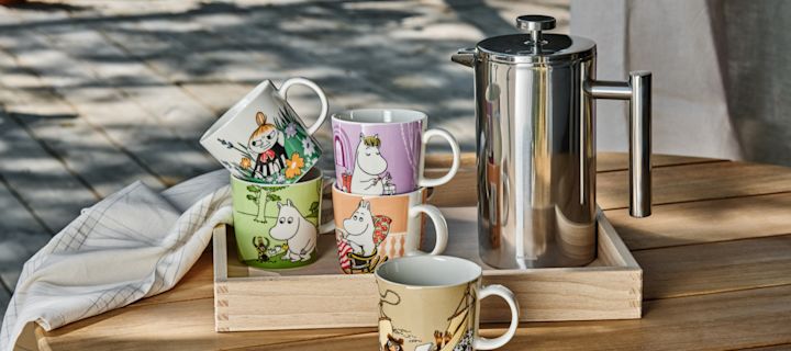 Flere ulike mummikopper fra Moomin Arabia står på et bord ute, ved siden av en presskanne.
