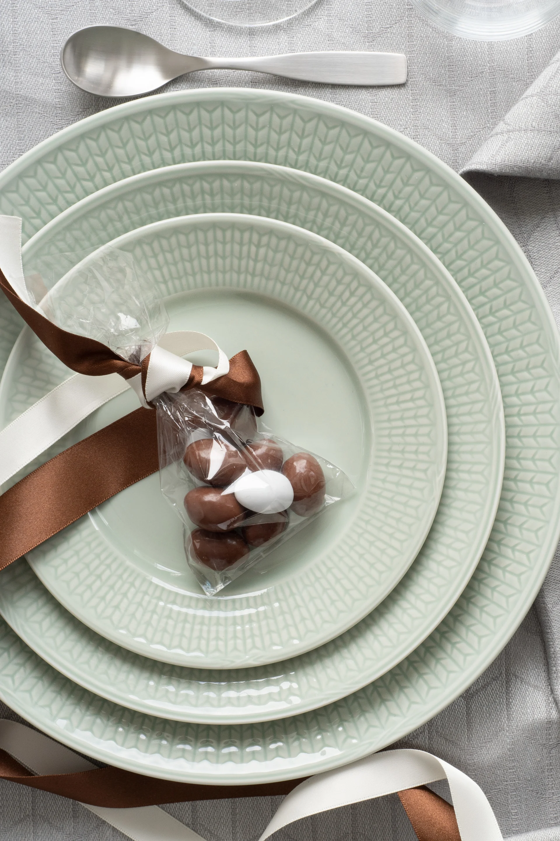 Stablede lysegrønne tallerkener med en gavepose sjokolade på en grå duk, ved siden av sølvbestikk.