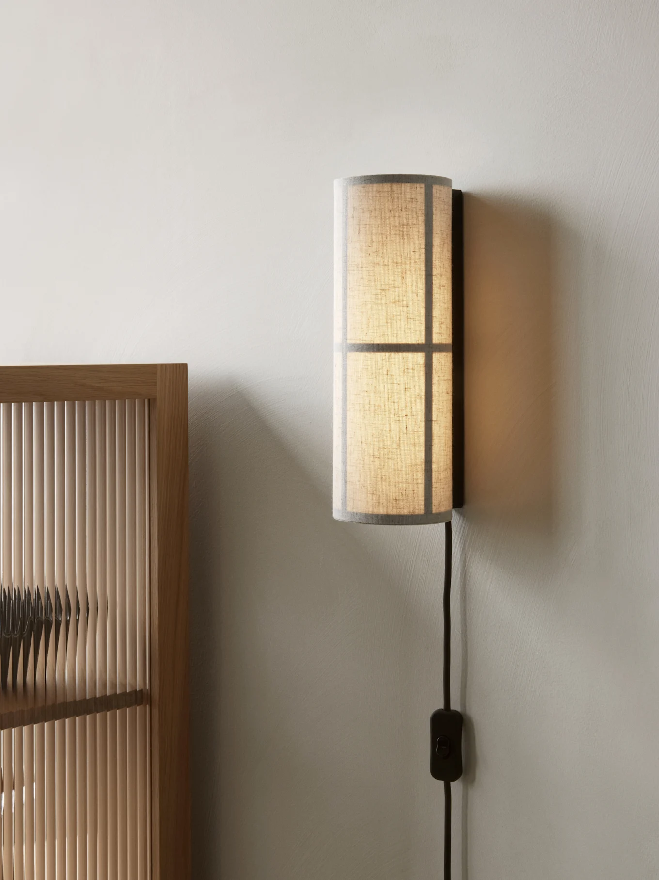 Sylinderformet vegglampe med en gjennomskinnelig stoffskjerm, montert på en lys vegg ved siden av en tresjalusiskjerm.