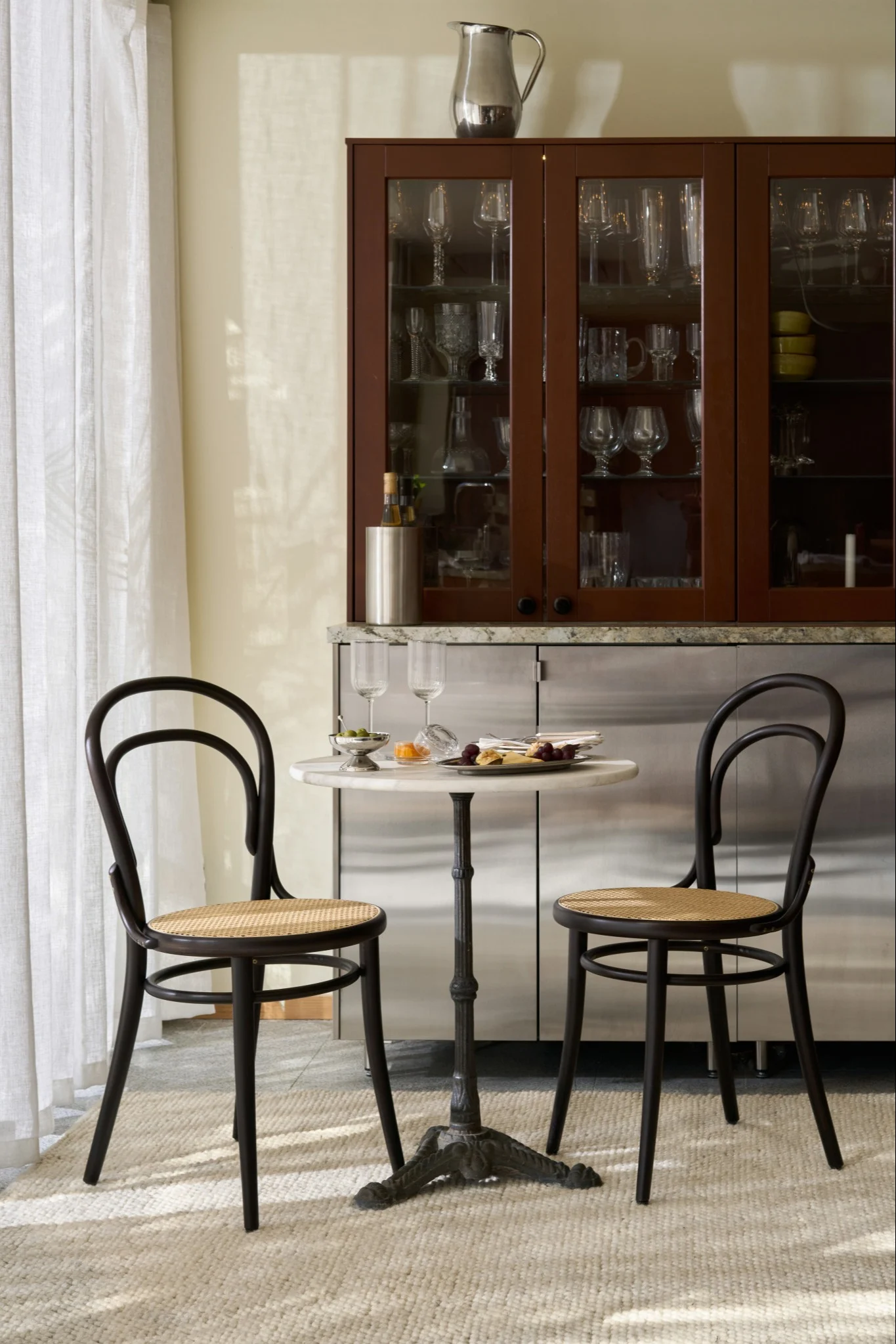 To mørke bentwood-stoler og et lite marmorbord med snacks foran et barskap med glassvarer.