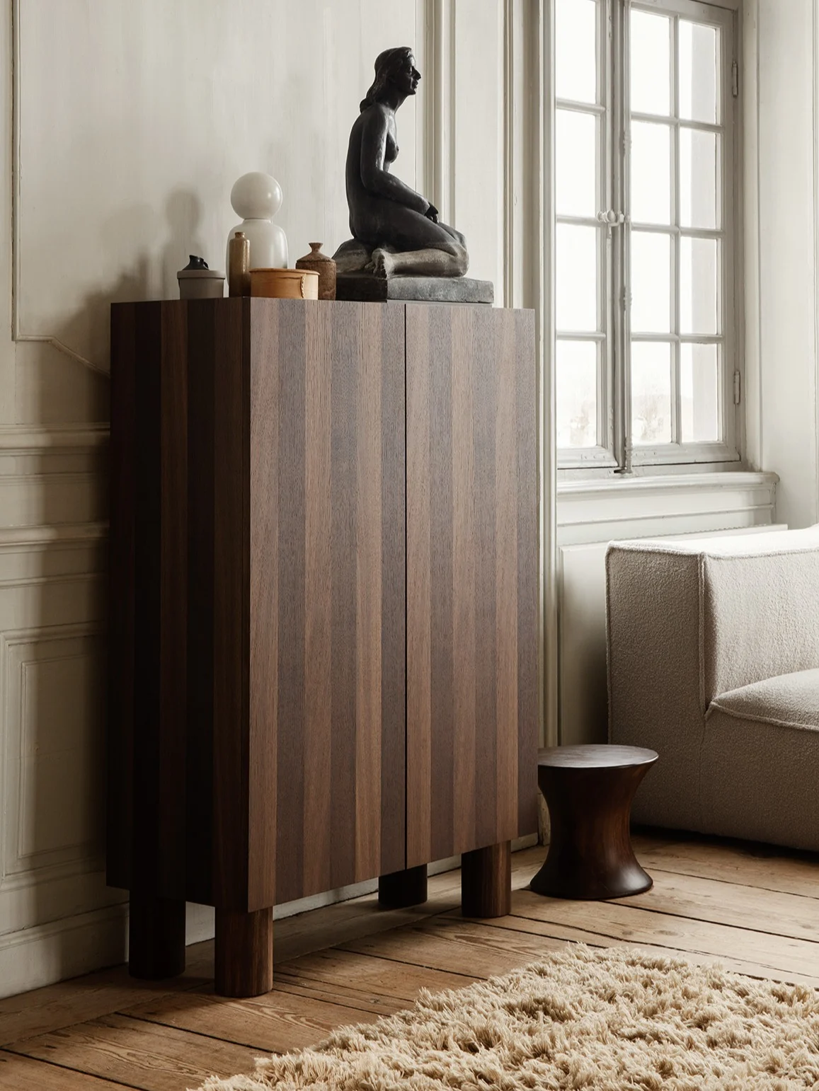Mørkt treskap med vertikale striper, toppet av en sittende skulptur, hvit lampe og dekor, ved siden av en teksturert sofa og mørk krakk.