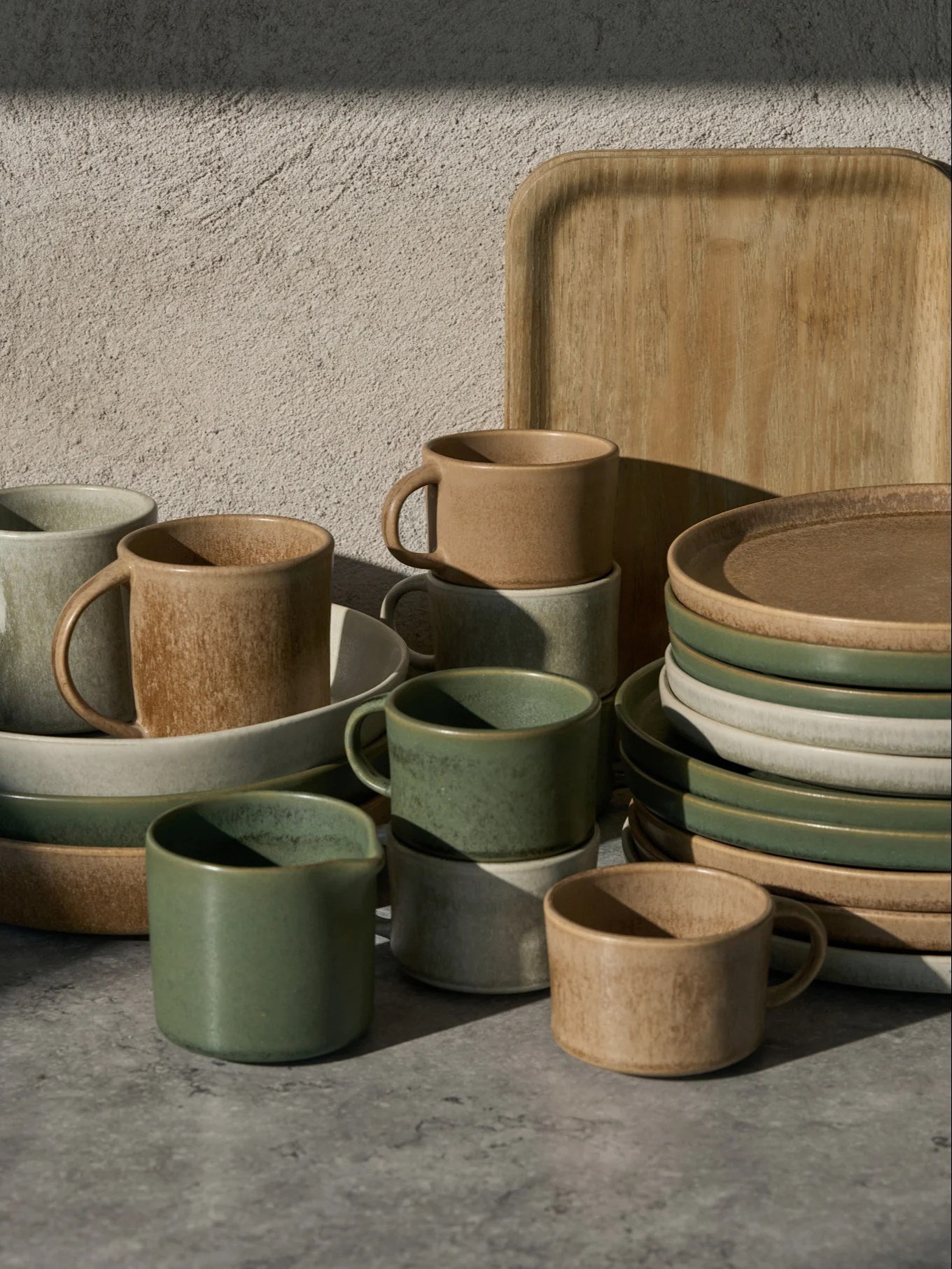 Samling av grønne, beige og lysegrå keramiske kopper, skåler og tallerkener, med et trebrett, på en grå overflate.
