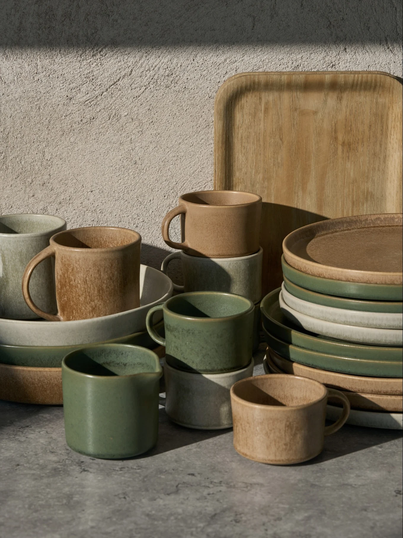 Samling av grønne, beige og lysegrå keramiske kopper, skåler og tallerkener, med et trebrett, på en grå overflate.