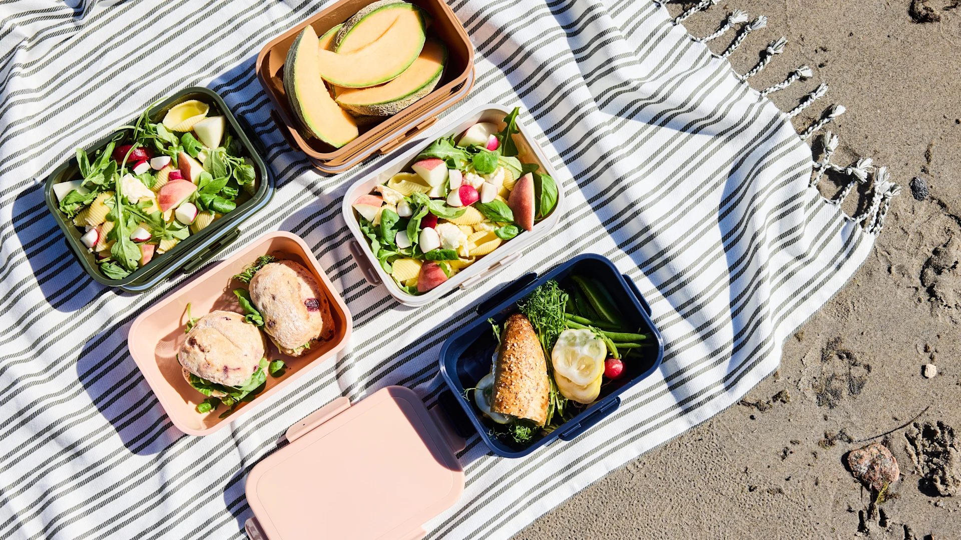 Piknik spredt på et stripete teppe på sand, med flere matbokser som inneholder frukt, pastasalat og smørbrød.