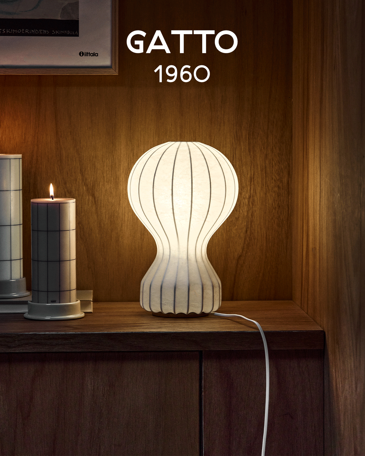 En glødende hvit riflet lampe med timeglassform står på en trehylle ved siden av et tent stearinlys. Tekst: GATTO 1960.
