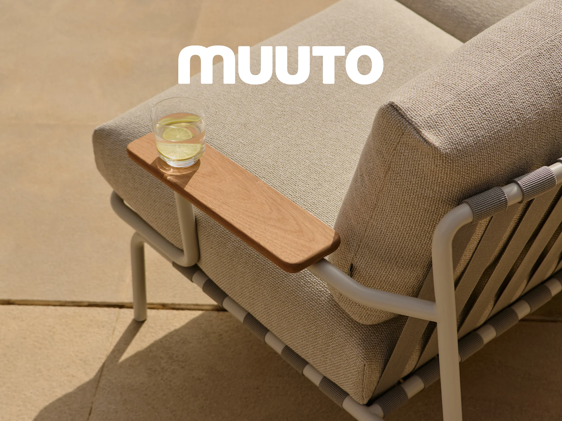 Nærbilde av en utendørs sofa med lyse stoffputer, et trearmlene med et glass vann og den hvite Muuto-logoen.
