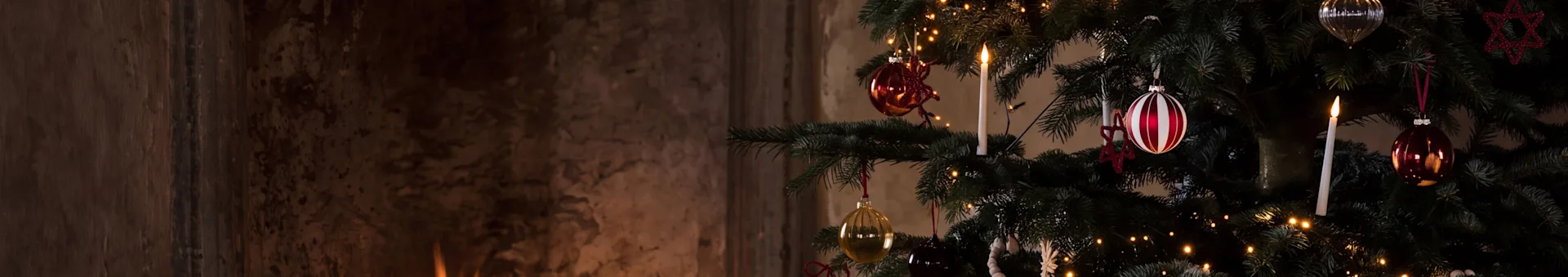 Et festlig juletre med ornamenter og et tent lys ved siden av en varm peis med brennende ved.