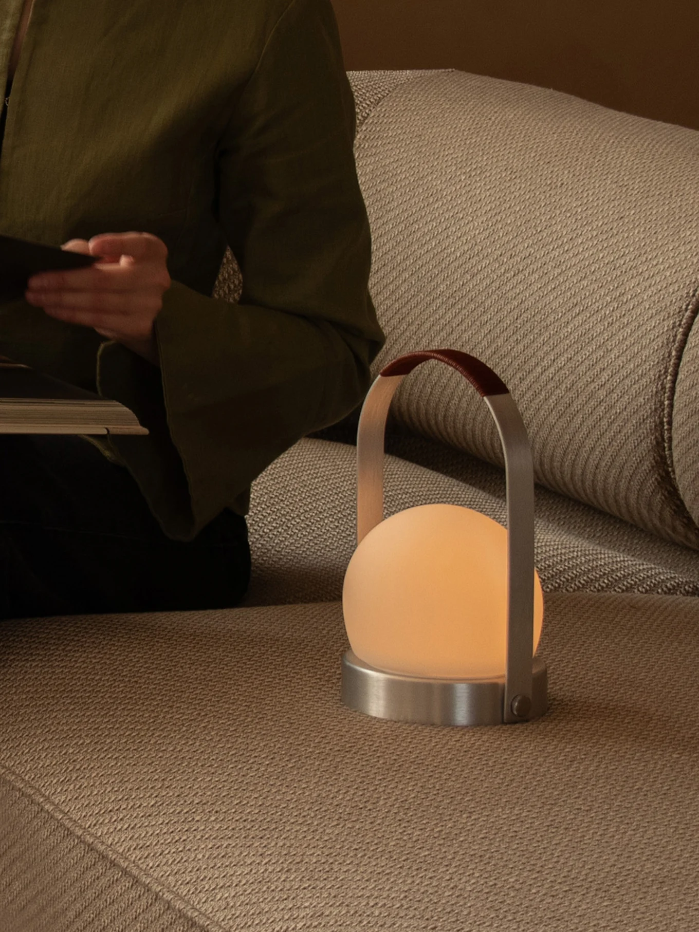 Person leser på sofa med en glødende bærbar lampe med frostet kule, metallfot og skinnhåndtak.