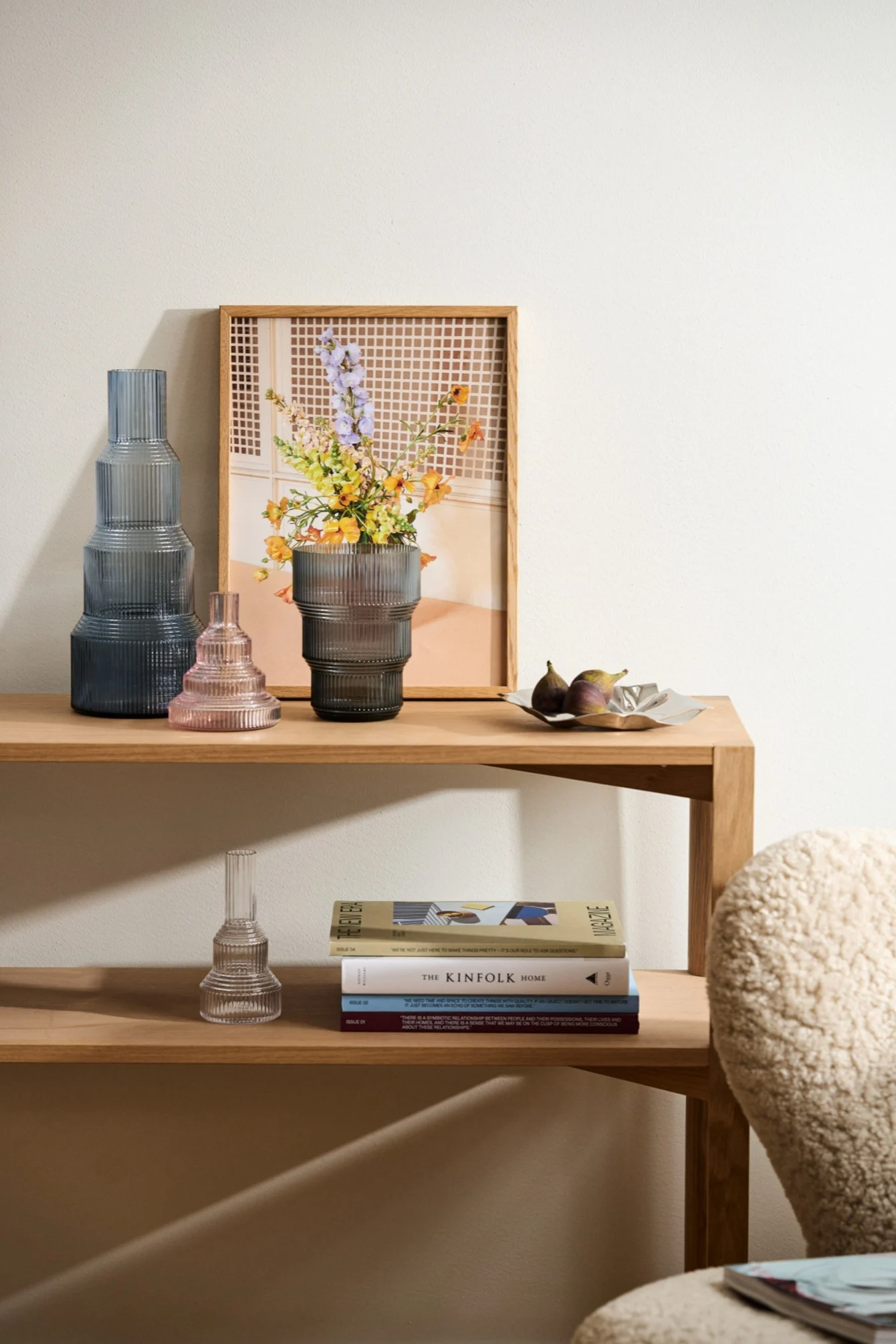 En trehylle med tre riflede glassvaser (høy mørkeblå, liten rosa, mellomgrå med blomster), et innrammet bilde, fiken og bøker.