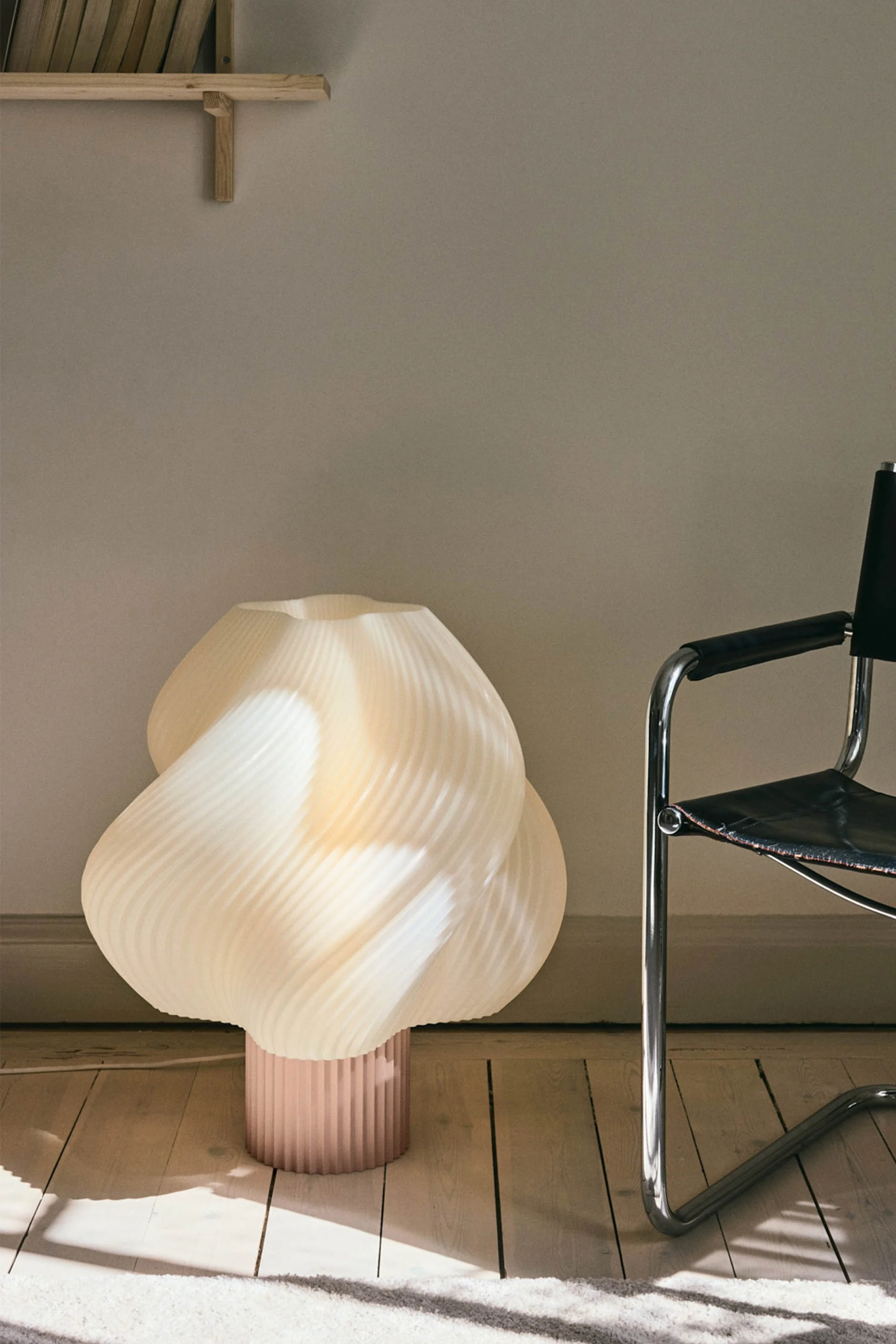 En skulpturell, plissert kremfarget gulvlampe med rosa base står på et tregulv ved siden av en sort skinn- og kromstol.
