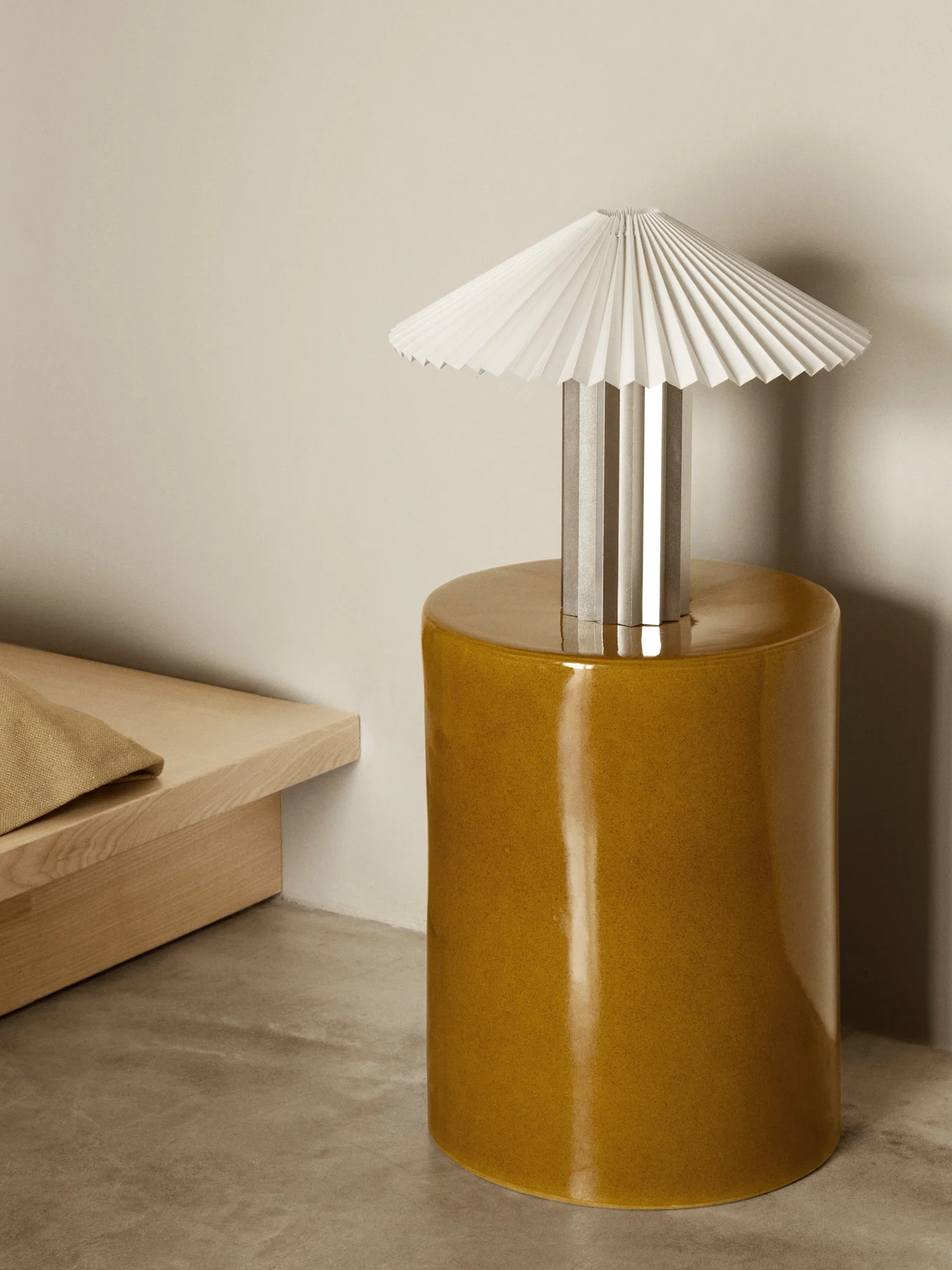 En bordlampe med hvit plissert skjerm og sølvfot står på et blankt sennepsgult sylindrisk sidebord i et minimalistisk rom.