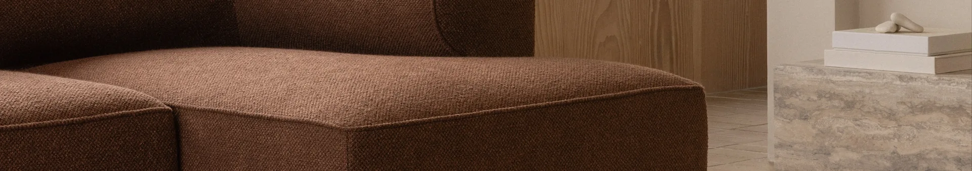 Nærbilde av en brun teksturert sofa, med en trevegg og dekorative gjenstander på en betongflate i bakgrunnen.