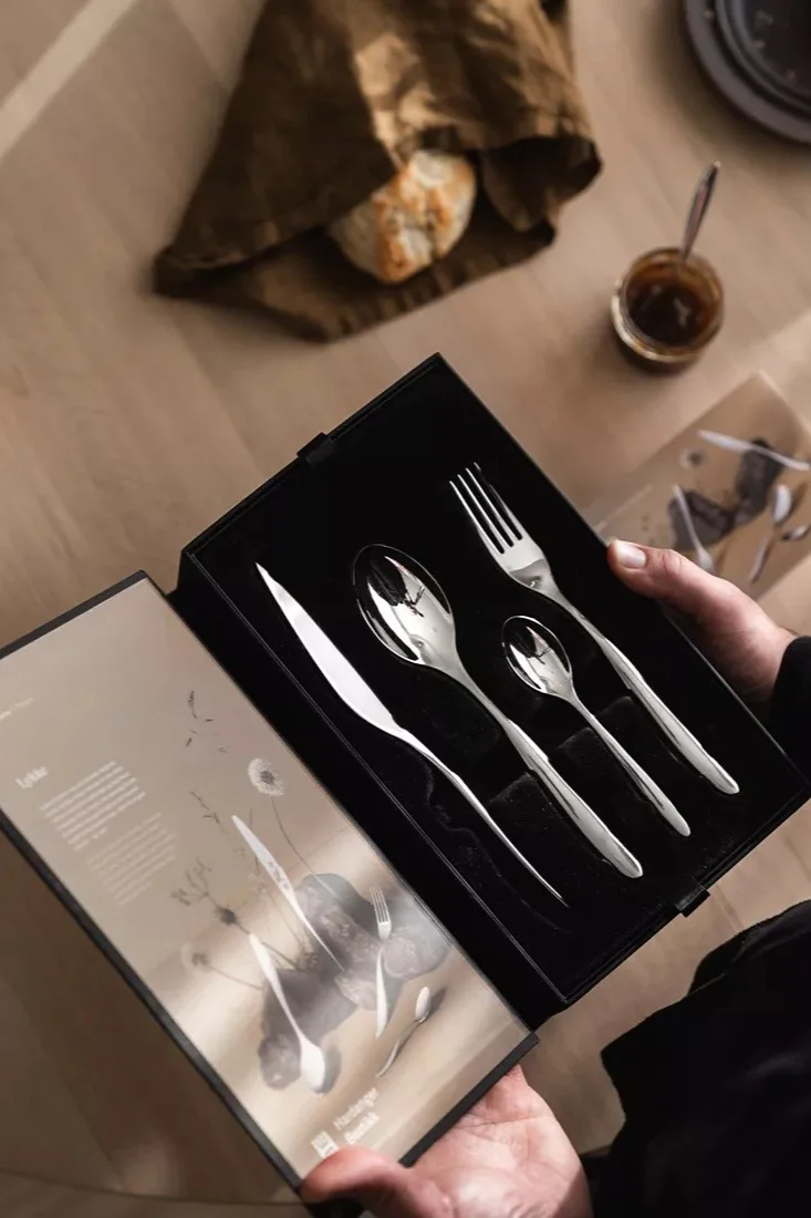 Hender holder åpen svart eske med et sett skinnende sølvbestikk: kniv, stor skje, liten skje og gaffel. Brød og syltetøy på et trebord.