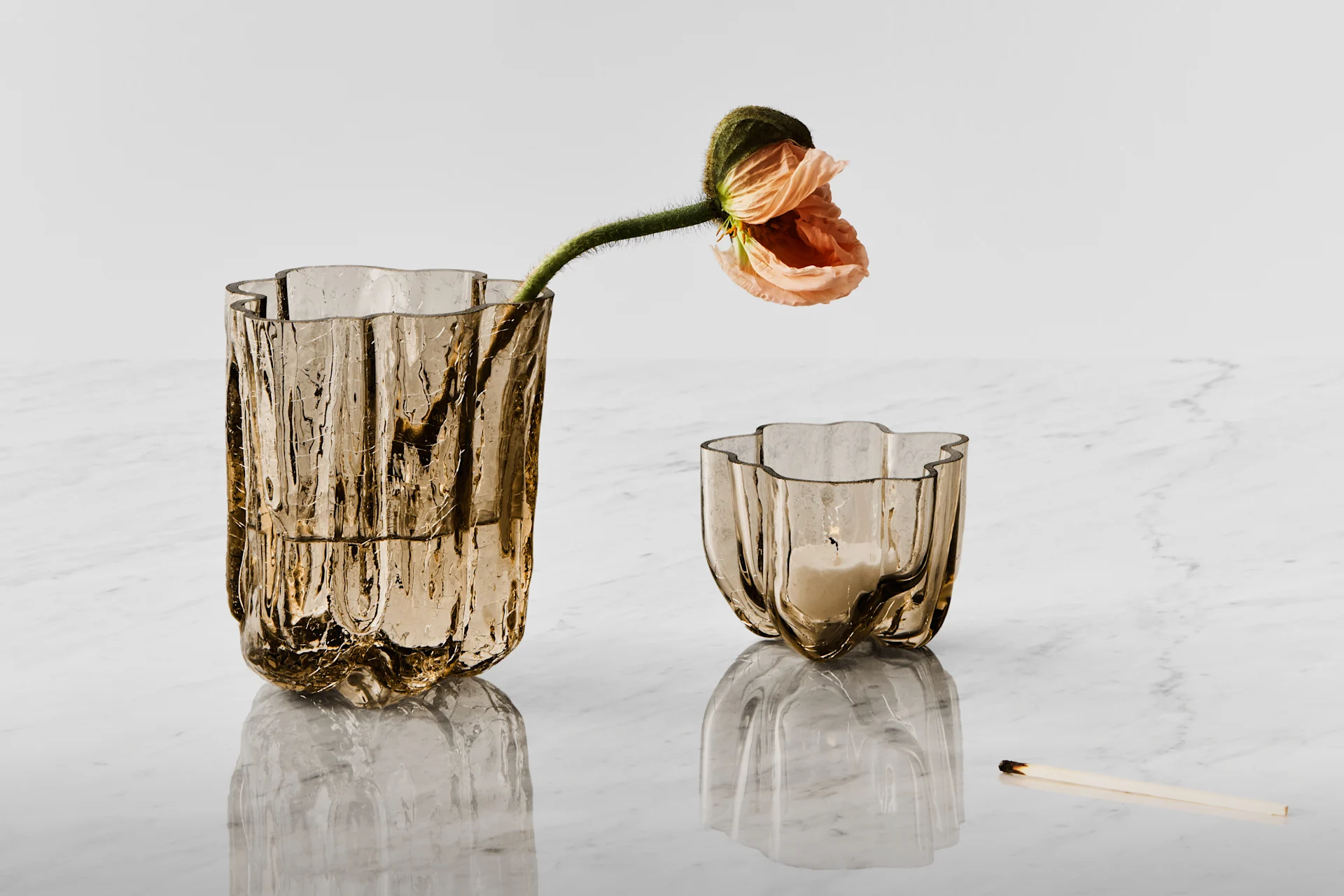 To teksturerte ravfargede glassbeholdere, den ene med en ferskenfarget valmue, den andre et tent lys, på hvit marmor ved siden av en brent fyrstikk.