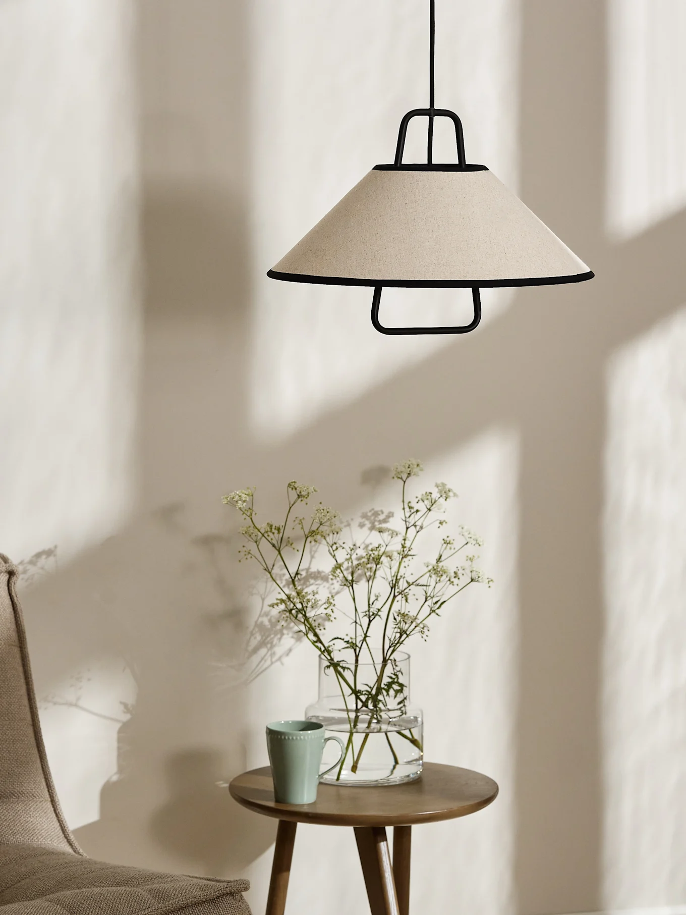 En konisk kremfarget pendellampe med svart kant henger over et sidebord i tre med en vase hvite blomster og et lysegrønt krus.