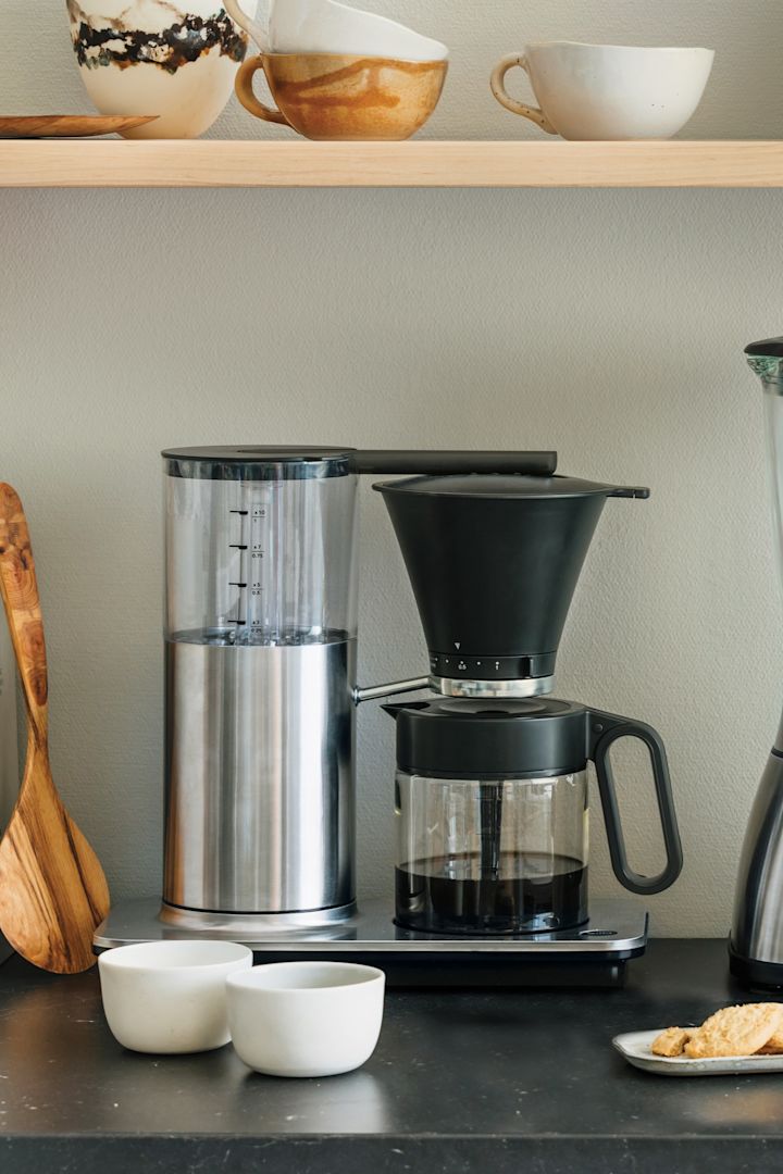 Du trenger også de riktige apparatene til kaffebaren din hjemme. Her kan du se en klassisk filterkaffemaskin fra Wilfa stående på en kjøkkenskjenk, med en hylle med boller og en brødrister over.