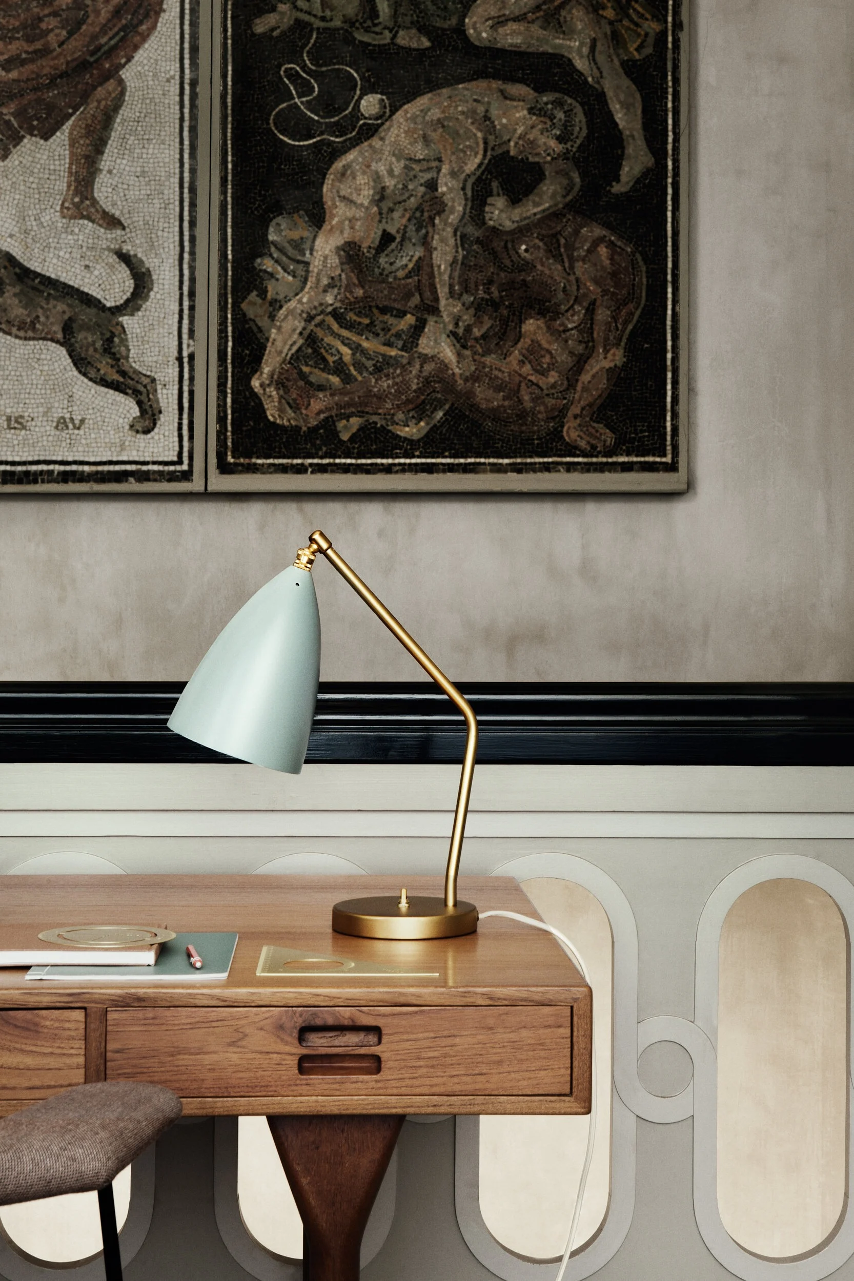 Her kan du se Grasshopper bordlampe i messing med en blågrå lampeskjerm som står på et skrivebord av tre.