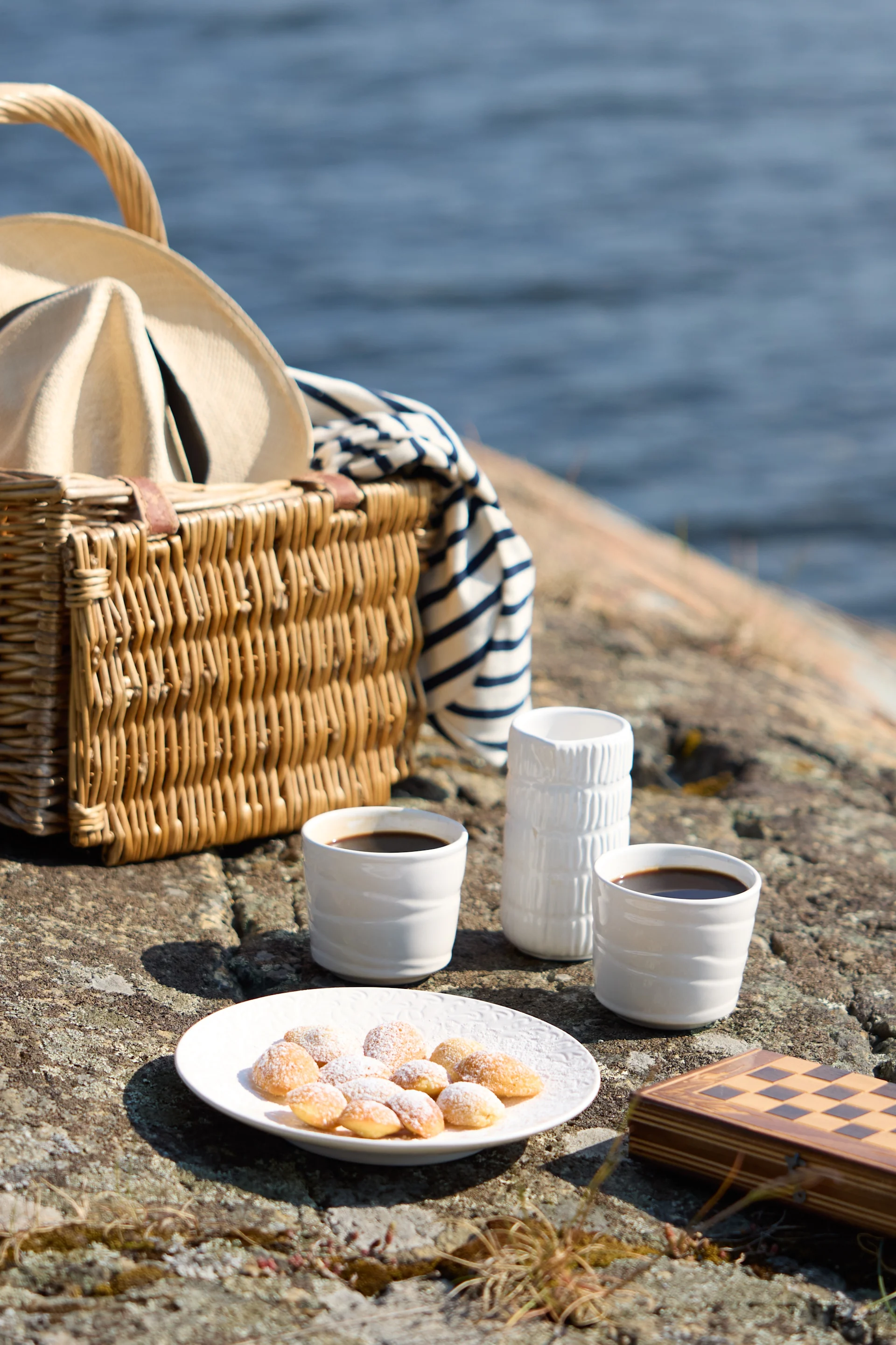 En piknik på klipper ved sjøen, med en flettet kurv med hatt og stripet teppe, to kopper kaffe, bakverk og et lukket sjakkbrett.