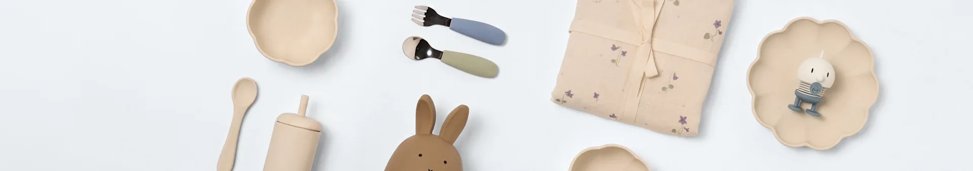 Babyartikler på hvitt: to kremfargede skåler, blått og grønt bestikk, blomstret stoff, kaninleke, treskje, beige kopp og en liten figur på en bølget tallerken.
