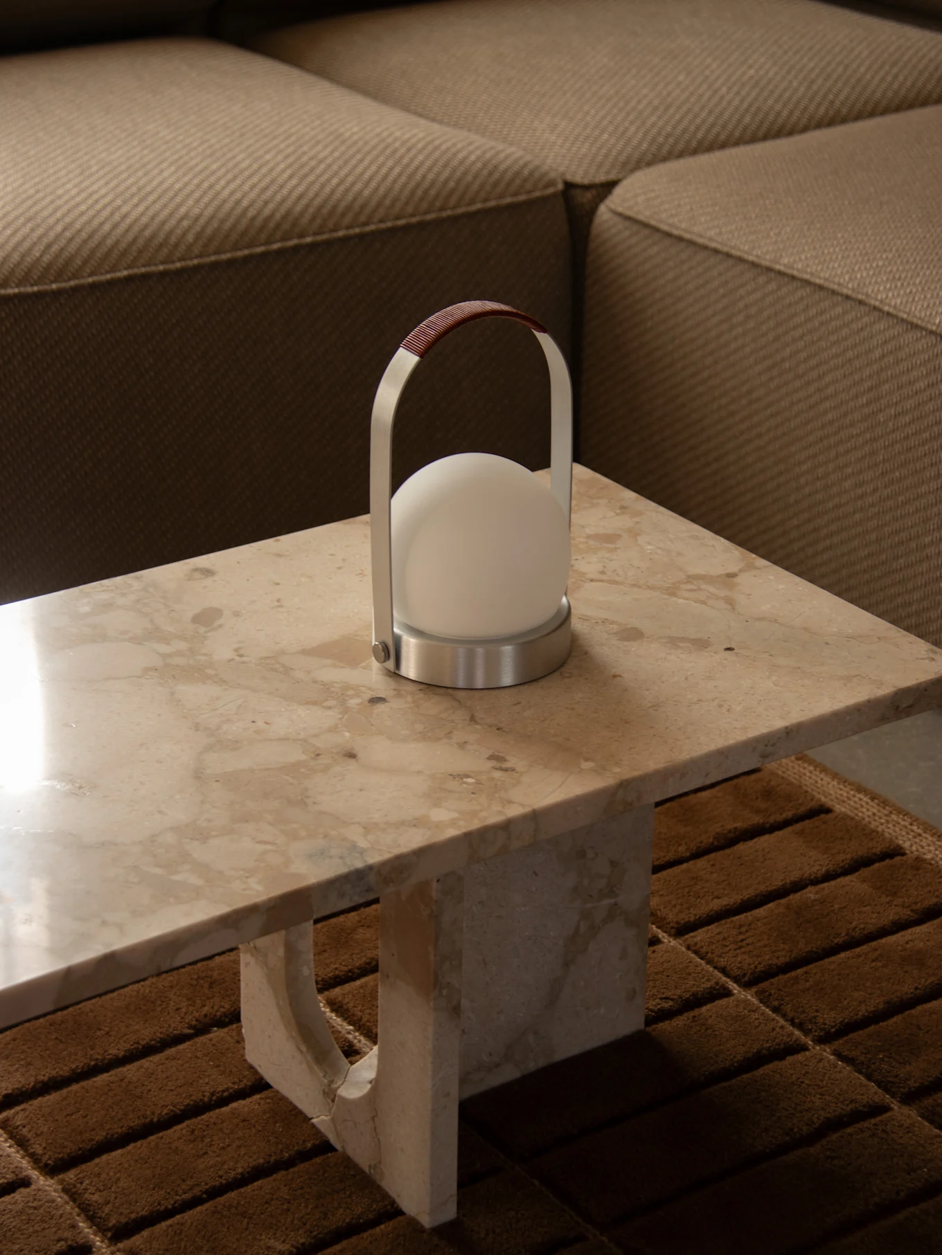 Moderne bærbar lampe med kuleformet hvitt lys, sølvramme og brunt lærhåndtak på et marmorbord ved siden av en sofa.