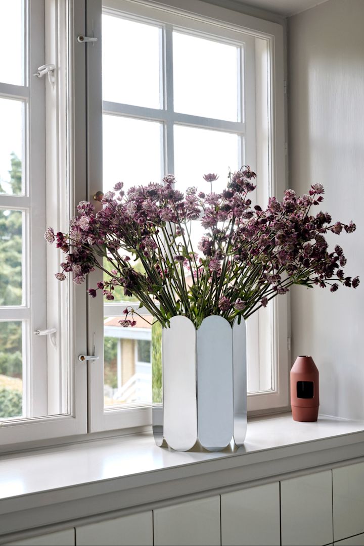 En pyntet vinduskarm med Arc-vasen og lilla blomster.
