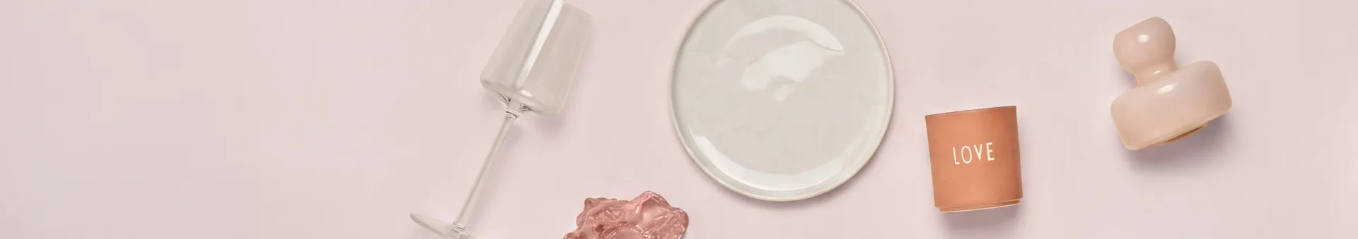 Oversikt over ulike dekorative gjenstander og servise på en lys rosa overflate. Fra venstre til høyre: et skråstilt klart glass, en kremfarget tallerken, en terrakotta "LOVE" lysestake og en rosa abstrakt skulptur. En mindre rosa bølgeformet gjenstand er under glasset.