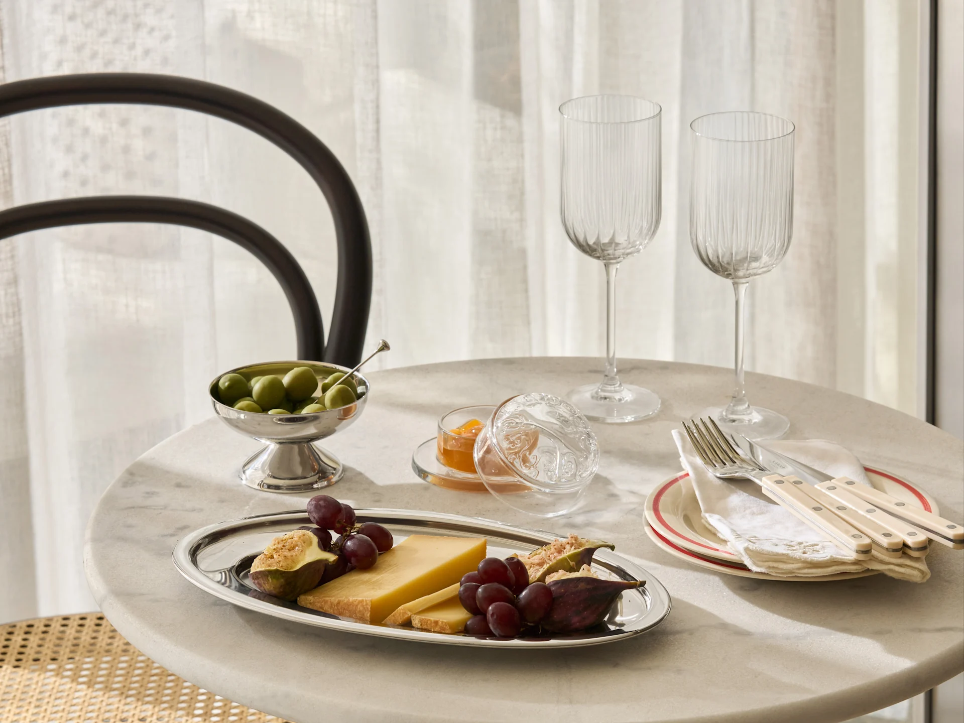 Et marmorbord dekket med et sølvfat med ost, druer, fiken, en bolle oliven, syltetøy, to riflede glass og bestikk på tallerkener.